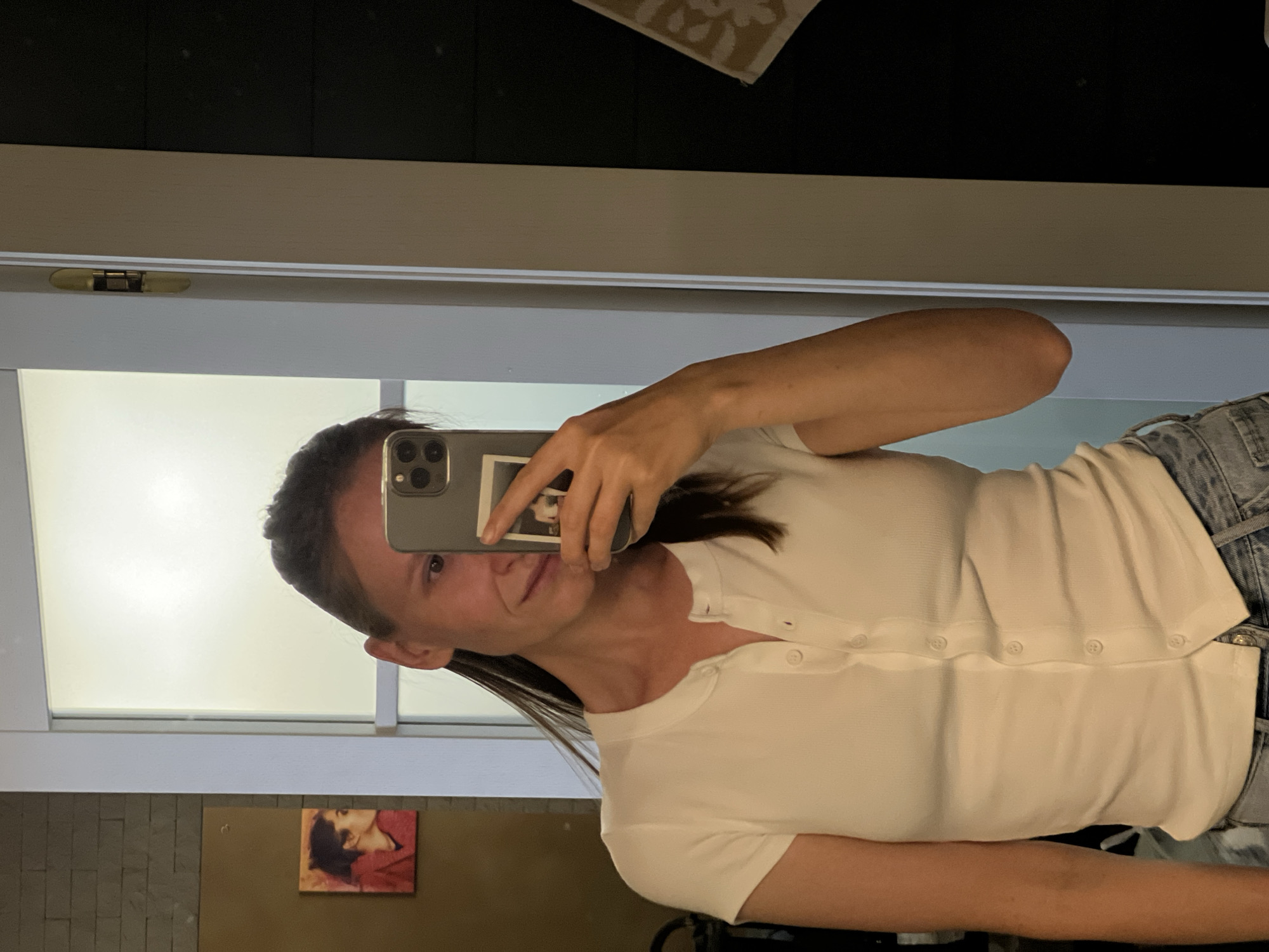A woman is taking a mirror selfie in a room. She is wearing a white button-up shirt and denim shorts. She is holding a phone in her right hand, which is angled to capture her reflection. In the background, there is a door, a mirror, and a small framed picture of a woman's profile. The environment appears to be a bathroom or dressing area. The location of Khimki, Russia is not depicted in the image.