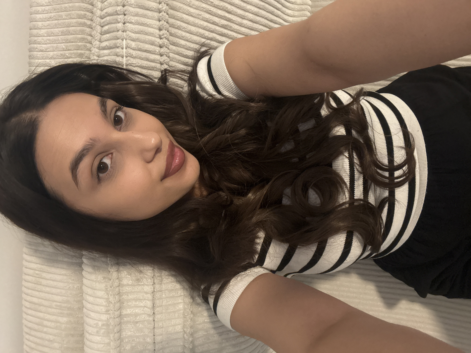 A woman with long, dark brown wavy hair, fair skin, and dark eyes is lying on a white, textured blanket or bedspread. She's wearing a white and black striped top with short sleeves and a dark pair of pants. Her arms are extended above her. The photograph is taken from a high angle, looking down at her. There is no indication or evidence to determine if this photo was taken in Mönchengladbach, Germany.