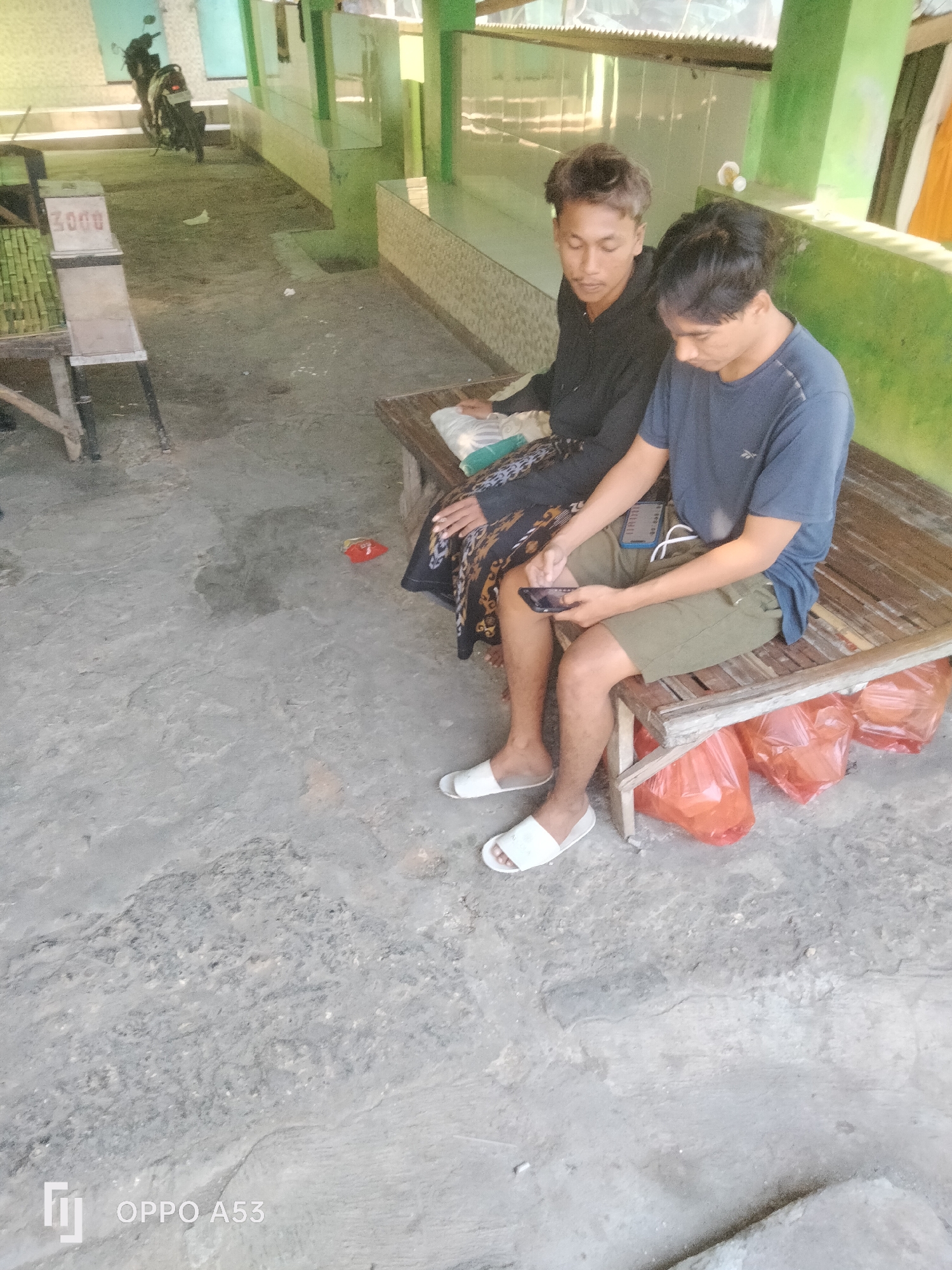 Two young men are sitting on a wooden bench in an outdoor area. The man on the left is wearing a black hooded jacket and a patterned sarong. The man on the right is wearing a blue t-shirt and green shorts. He is holding a smartphone in his right hand and looking down at it. Another smartphone is resting on the bench next to him. Underneath the bench, there are three large orange plastic bags. In the background, there is a motorcycle parked on steps and a green wall with tiled accents. The ground is paved with concrete. The overall scene appears to be in a public space in Badung, Indonesia, possibly a marketplace or a rest area.