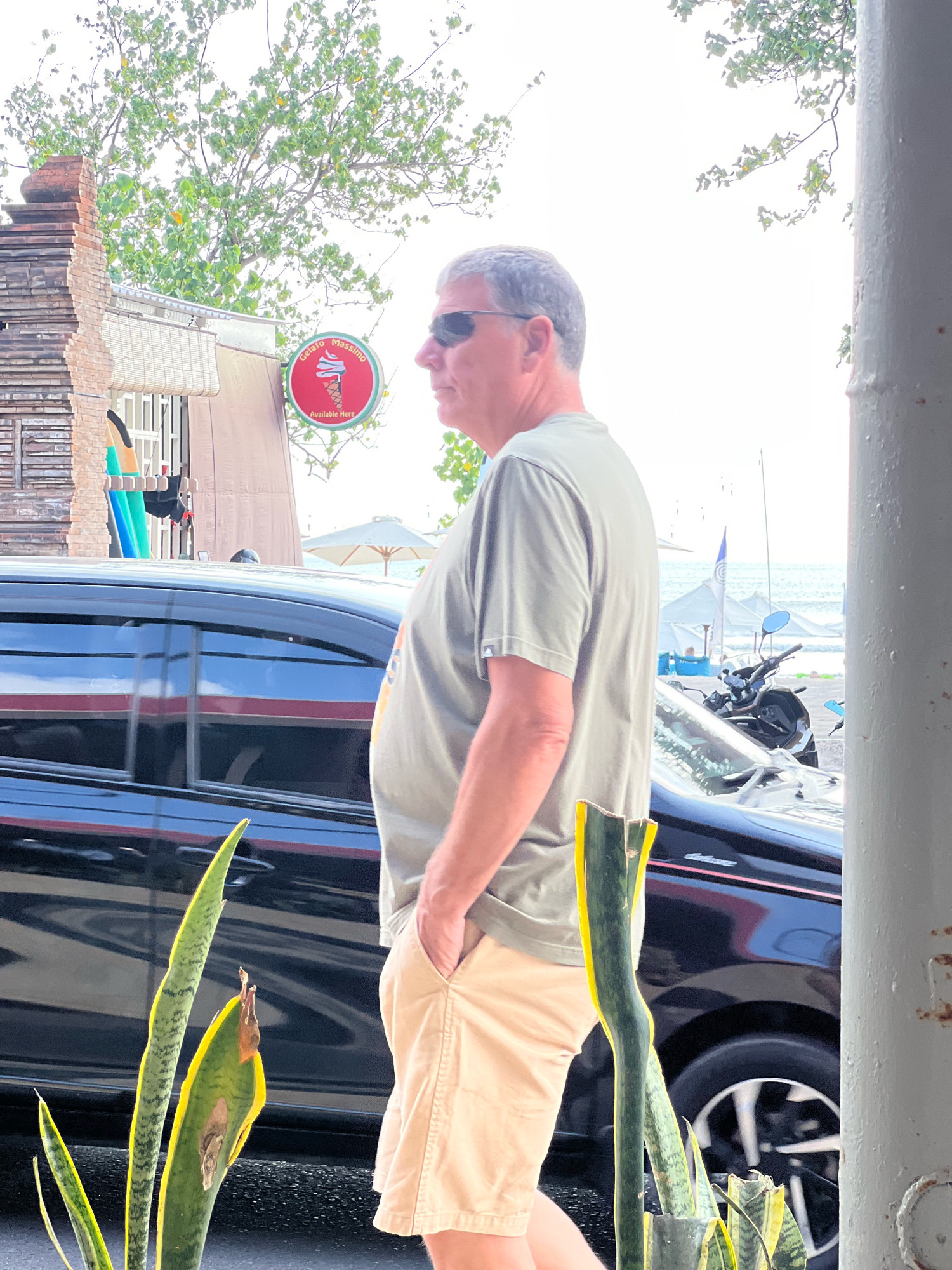The image depicts a man in his 50s, with graying hair and sunglasses, standing outdoors in Kuta, Indonesia. He is wearing a light olive green t-shirt and light beige shorts, with his right hand in his pocket. The man is positioned in the foreground, facing right and looking away from the camera.
Behind him, a black car is parked. To the left of the car, there is a building with a brick facade and a sign that says "Gelato Massimo" with an image of an ice cream cone. In the background, beyond the car, a beach is visible with several white umbrellas and a motorcycle parked on the sand. Trees with green foliage are also present in the scene. The overall atmosphere suggests a warm, sunny day in a coastal town.