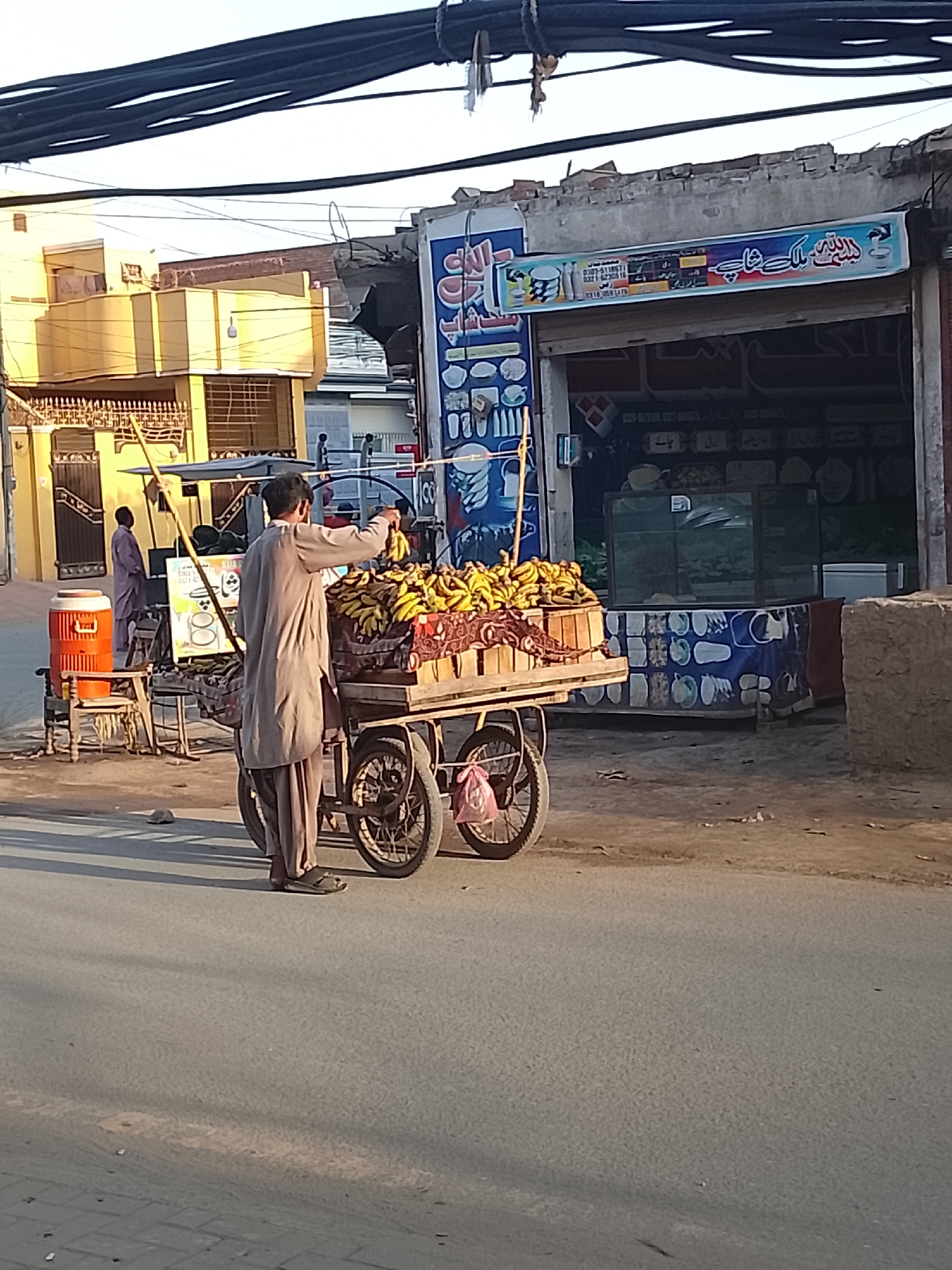 The image captures a street scene, likely in South Asia, characterized by a fruit vendor and urban architecture. In the foreground, a man wearing a light brown shalwar kameez and flip-flops stands behind a wooden fruit cart laden with numerous bunches of ripe yellow bananas. He appears to be arranging the fruit. The cart has two large rear wheels and a smaller front wheel, suggesting it might be a modified bicycle or tricycle cart.
Behind the fruit vendor, to the left, is a vibrant yellow multi-story building with intricate metalwork fences and barred windows, possibly a residential structure. Further back and to the right, a second man in a darker traditional outfit stands near another smaller cart or stand, which has a red and orange water cooler on it.
To the right of the main fruit cart is a shop or building with a partially open storefront. A prominent blue and white signboard above it displays text in Urdu/Arabic script, which reads "رِشْتَه مل کِ شاپ" (Rishta Milk Shop, or a similar name) and lists three phone numbers: 0323-5118571, 0321-6230516, and 0316-0591476. The signboard also features images of cups and bowls, suggesting the shop might sell kitchenware or serve beverages. A blue printed cloth with white circular patterns covers the lower display area of the shop.
The scene takes place on an asphalt road, with paved sections visible on the lower left. Long shadows cast on the ground suggest either early morning or late afternoon. Thick black electrical or utility cables stretch across the top of the frame, indicating an urban setting. The sky is bright and clear. The overall impression is a bustling yet currently quiet commercial street.