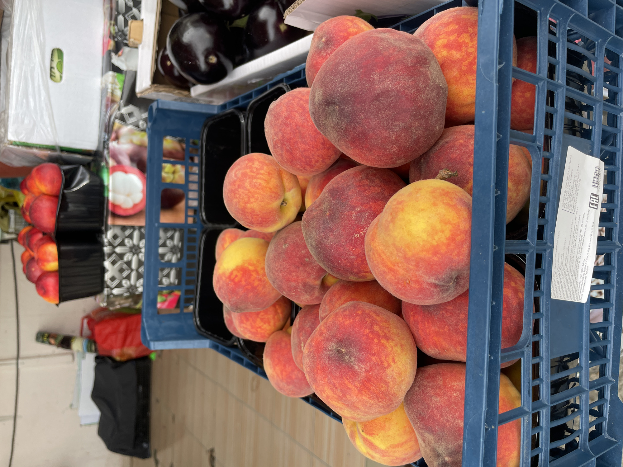 A blue plastic crate is filled to overflowing with fuzzy peaches. Another crate containing eggplants is visible to the upper left of the peach crate, and another appears to hold mangosteen. A white box is visible at the top left corner. The crate and box labels include the words “TURKIYE” and “Россия г. Сочи ул.” which suggest the location is in Sochi, Russia. The full address visible is Россия г. Сочи ул. Ров 40А, пом. 10. 11. 12, 1234.