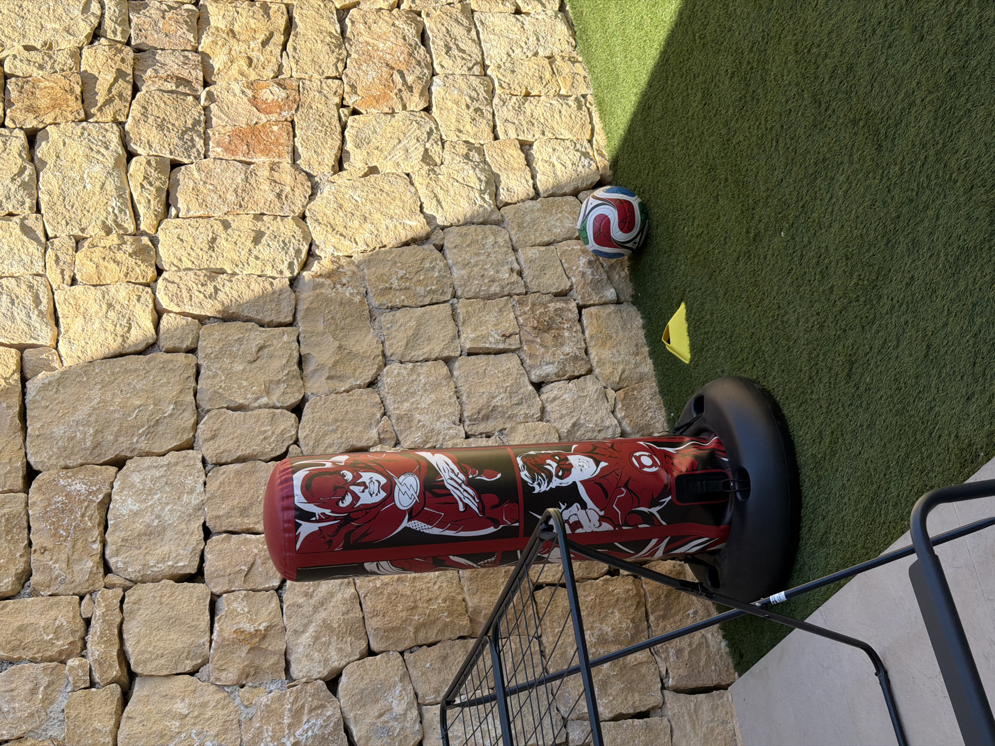 The image depicts an outdoor scene with a dry-stone wall constructed from large, light-brown, irregular blocks as the backdrop. In the foreground, artificial green turf covers the ground. To the left, a black metal drying rack is positioned partially on a light-colored stone patio. In the center, an inflatable punching bag decorated with comic book characters, primarily "The Flash" in red and white, stands on the turf. Next to the punching bag, a soccer ball with a red, white, and blue design rests on the turf. A bright yellow traffic cone is also visible on the turf to the left of the soccer ball. The scene is illuminated by sunlight, casting shadows on the wall and turf. The location is identified as Finestrat, Spain.