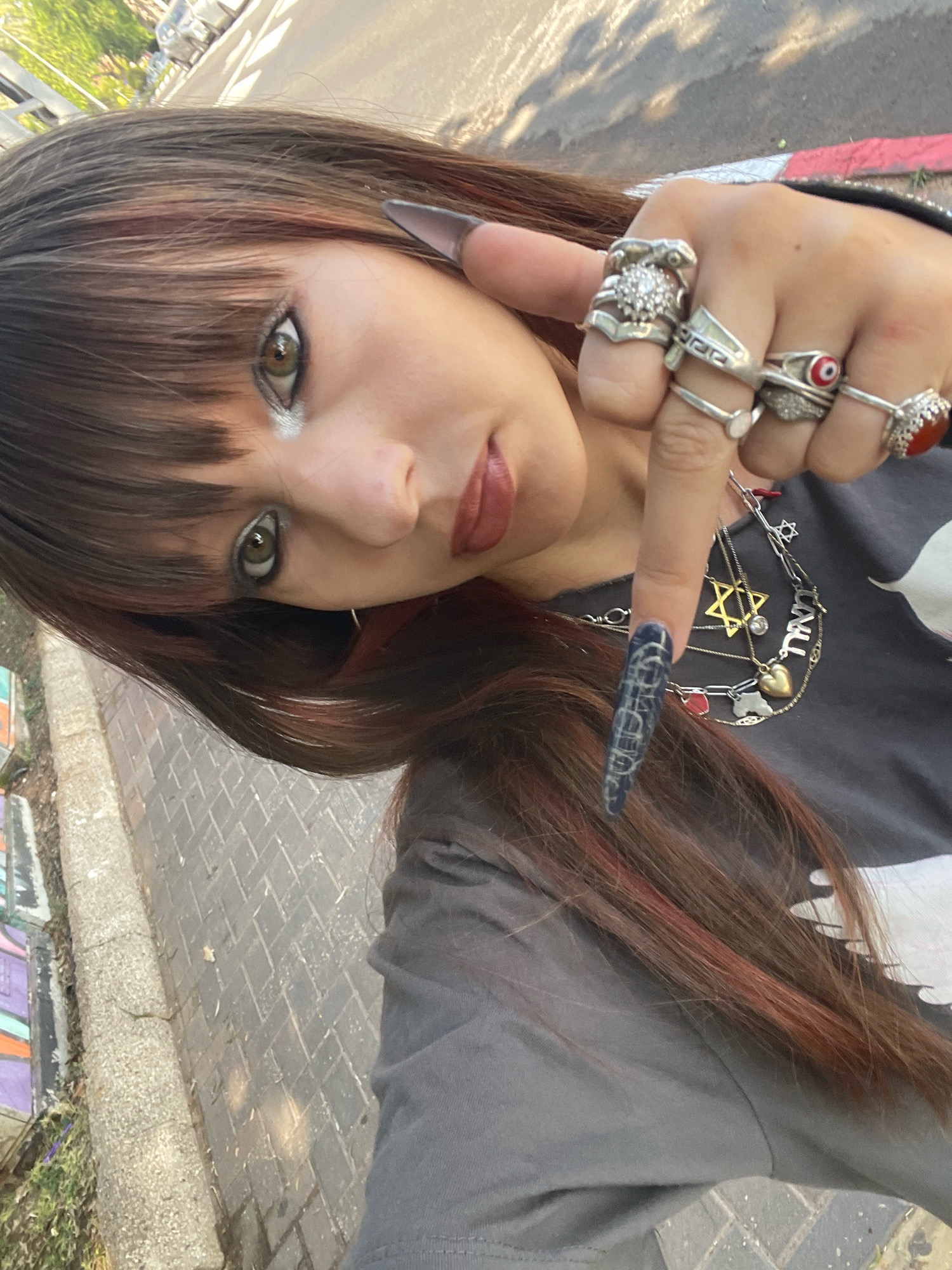 A young woman with brown hair and bangs, some strands of which are dyed reddish-brown, is taking a selfie outdoors. She is wearing a dark gray t-shirt and multiple silver rings on her right hand, including one with a red stone and another with an evil eye motif. Her fingernails are long and painted with a dark blue and white pattern. She has a necklace with several charms, including a Star of David, and what appears to be Hebrew lettering. Her makeup includes dramatic eyeliner and dark lipstick. She is standing on a sidewalk made of brick pavers, with a curb and some patchy grass and graffiti to her left. In the background, a street is visible with parked cars and trees. The overall lighting suggests it is daytime. The scene is consistent with an outdoor urban environment, possibly in Raanana, Israel, as indicated by the presence of Hebrew lettering on her necklace and the general appearance of the street and sidewalk.