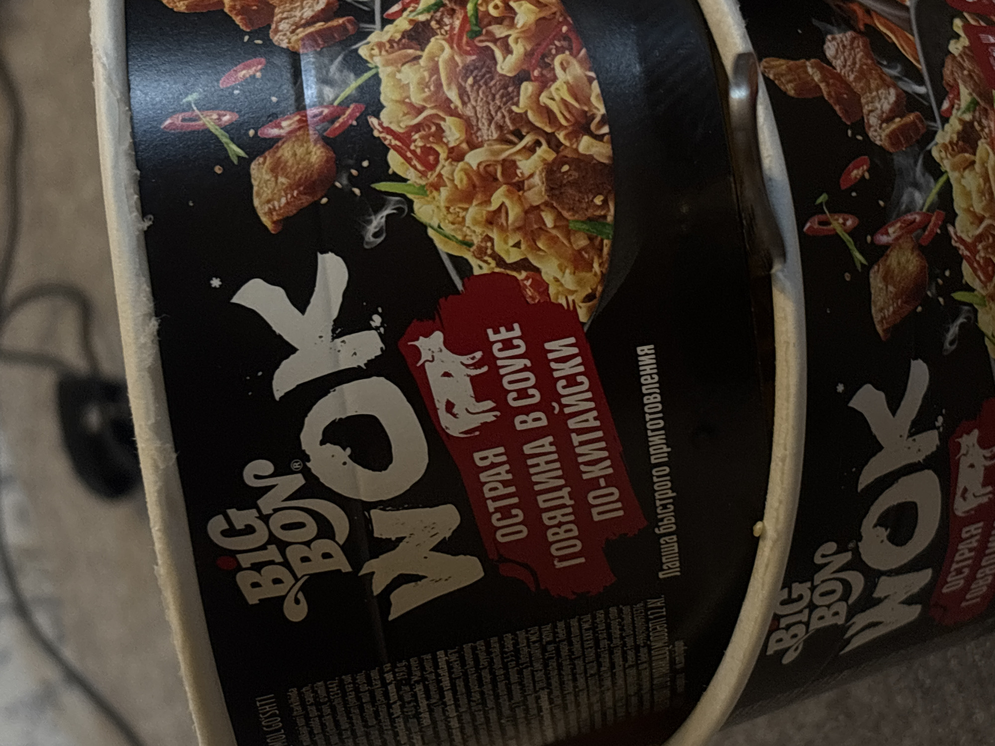The image shows two containers of "Big Bon WOK" instant noodles, featuring a black background with images of beef and noodles. The product name is prominently displayed in large, stylized white letters. Below, in red, is the Russian text "ОСТРАЯ ГОВЯДИНА В СОУСЕ ПО-КИТАЙСКИ," translating to "Spicy Beef in Chinese Sauce." Additional Russian text indicates it is instant noodles.
There is no indication of the location being Gyumri, Armenia.
