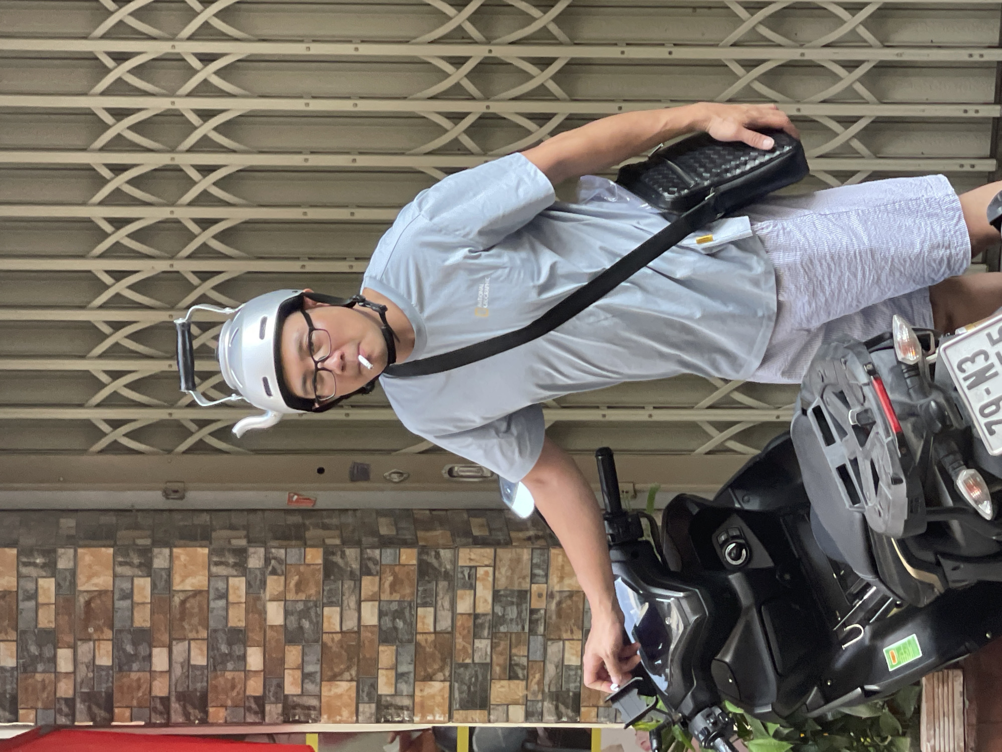 A man is sitting on a parked black scooter, smoking a cigarette. He wears a gray t-shirt with "National Geographic" printed on it, striped shorts, glasses, and a silver helmet. He has a black bag slung across his body. The scooter has a license plate with "79-N3" visible. The background includes a gray rolling metal gate and a brown and gray tiled wall. The location is Nha Trang Ward, Vietnam.