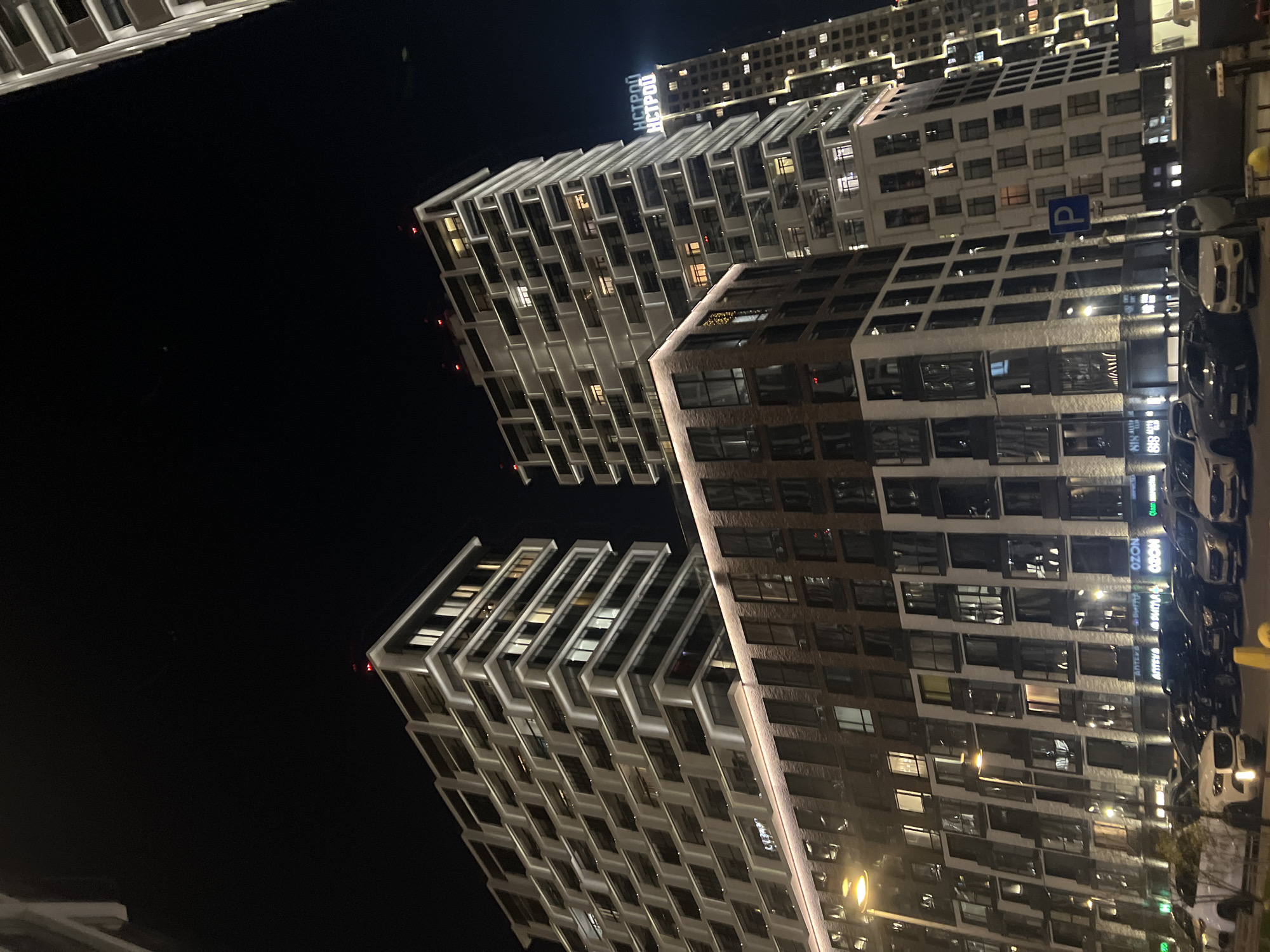 The image shows a nighttime view of modern buildings in Moscow, Russia. The buildings are characterized by their contemporary design with repetitive window patterns and projecting balconies. The façade colors vary between light beige and dark brown. The buildings appear to be apartment complexes. A parking sign is visible near the buildings. Cars are parked along the street in front of one of the buildings. The overall environment suggests an urban residential area with illuminated windows and streetlights. The skyline is dark, and the city lights provide the primary source of illumination.