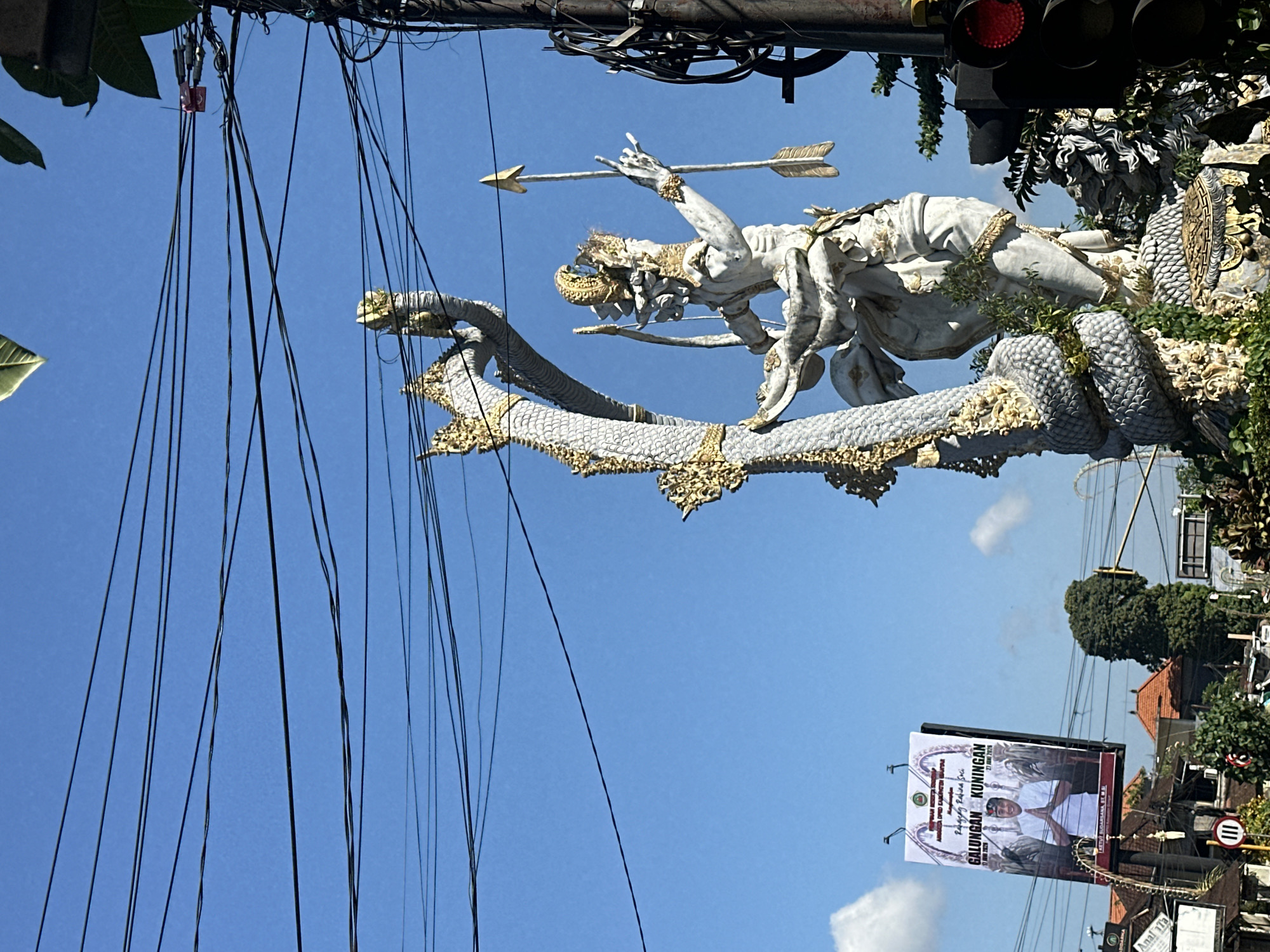 The image shows a statue of a figure, possibly a deity or mythical character, wrapped in a serpent-like creature. The statue is white with gold accents and appears to be made of stone or concrete. The figure is holding a bow and arrow, with an arrow nocked and ready to be fired. The serpent also has gold accents and is intricately detailed. The statue is set against a bright blue sky with some scattered clouds. In the background, there are power lines crisscrossing the sky, a billboard with text and an image of a man, and some trees and buildings. A traffic light with a red light illuminated is visible on the right side of the image. The overall impression is of a vibrant, possibly religious or cultural landmark. The location is indicated to be in the city of Ubud District, Indonesia.
