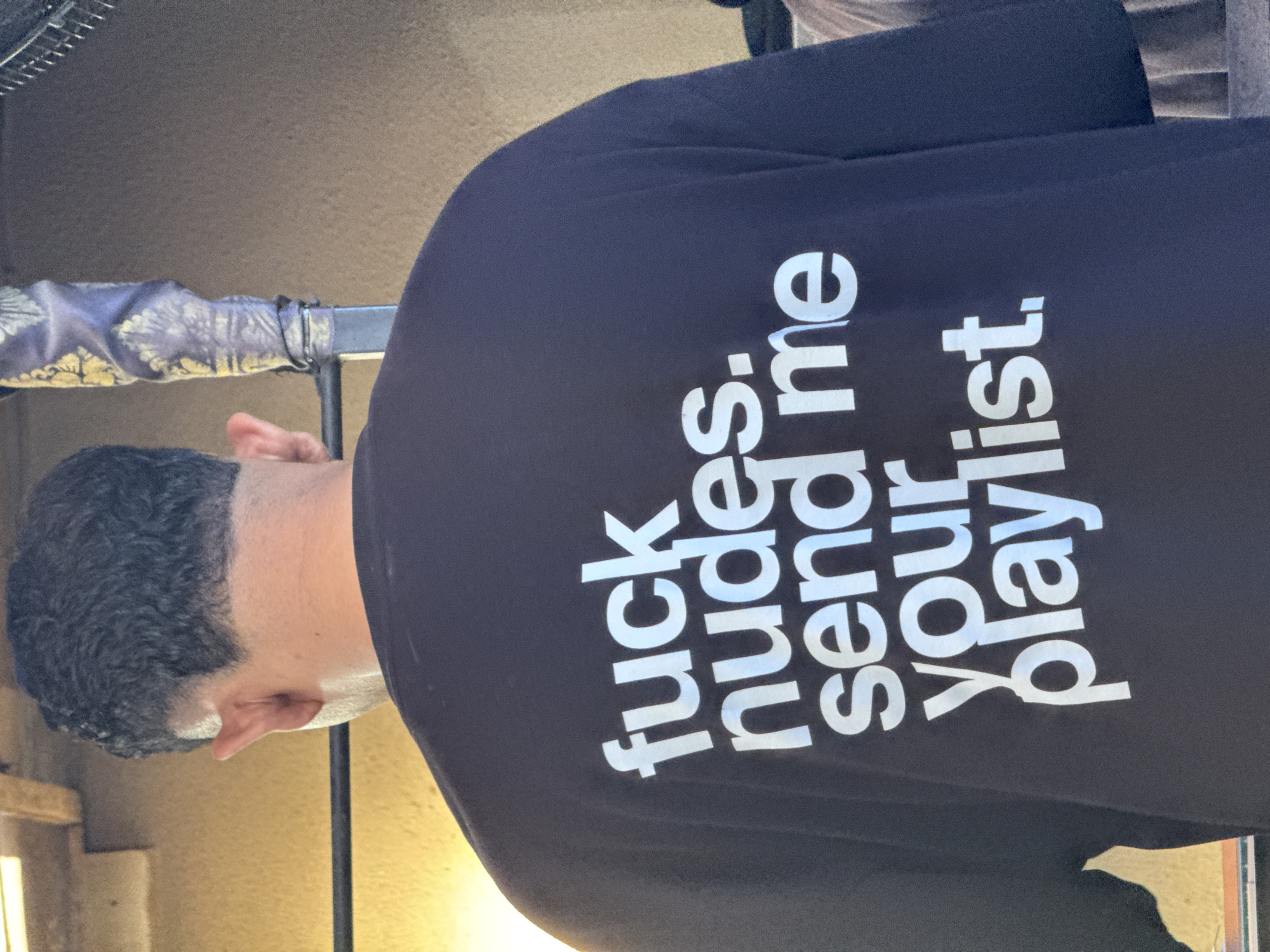 A man is wearing a black shirt with the text "fuck nudes. send me your playlist." in white. He is indoors, near a metal structure. The location is not evident from the image.