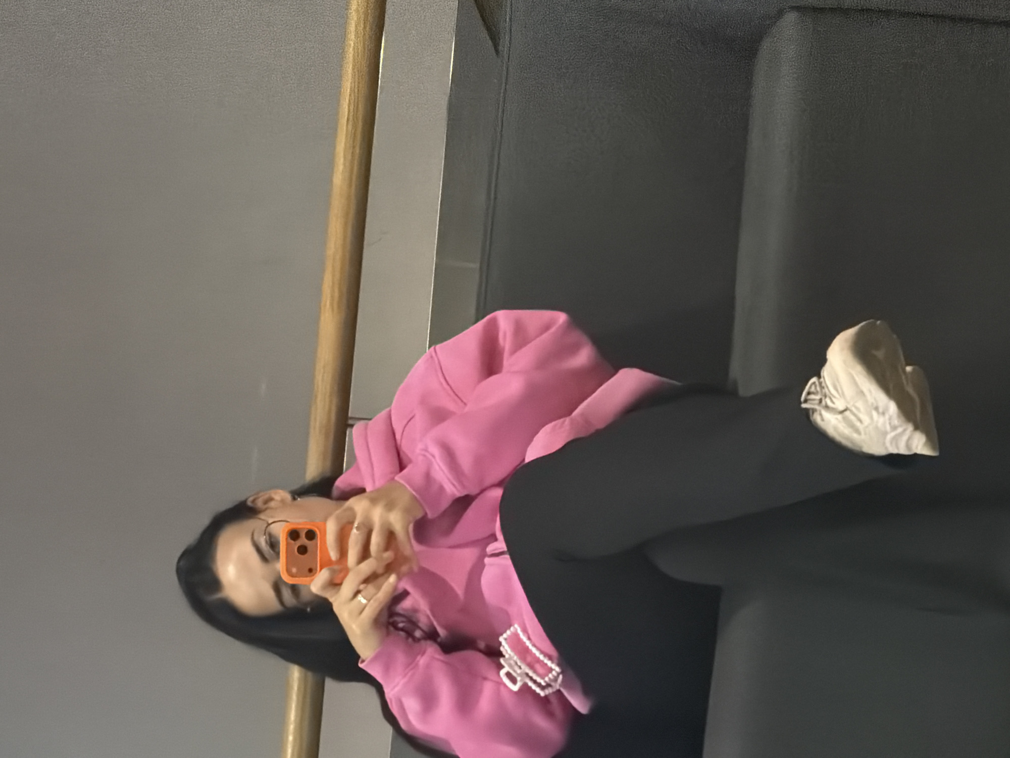 A woman lies on a dark gray surface, presumably a sofa, wearing a pink hoodie and black leggings. She is holding an orange smartphone, seemingly taking a photo of herself. She also wears white sneakers. The background consists of a light gray wall and a wooden railing. It is impossible to determine the location as Saint Petersburg, Russia, based solely on the given image.