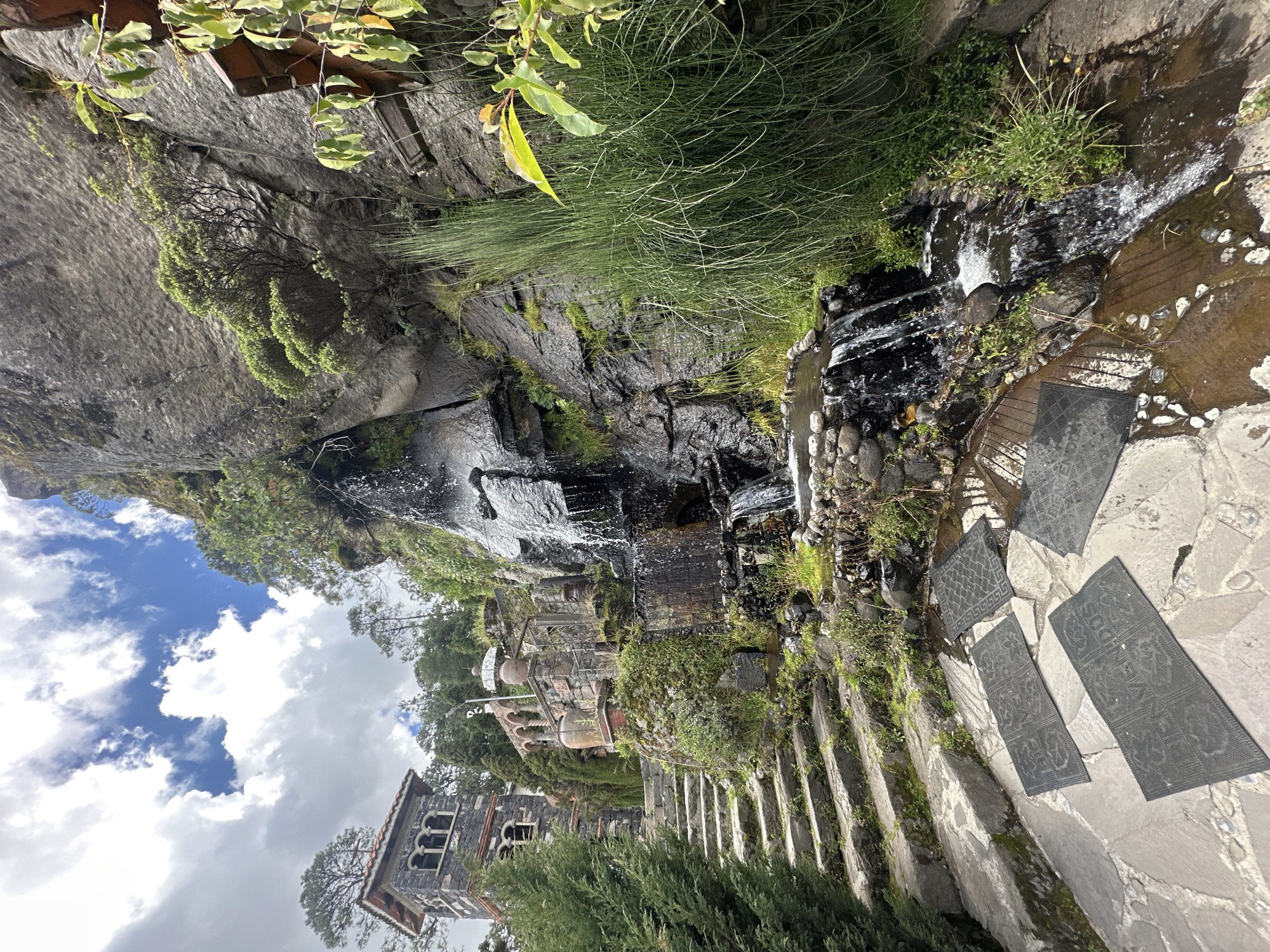 In the city of San Mateo Ozolco, Mexico, a multi-tiered stone staircase ascends towards a structure resembling a tower, built from dark stone with arched windows and a red-tiled roof. To the right of the staircase, a natural waterfall cascades down a rocky cliff face, feeding into a small, shallow pool at the base. Lush green vegetation, including tall, reedy plants and mossy patches, grows around the waterfall and along the rocks. The ground in the foreground is paved with irregular flagstones, and several dark-colored mats with geometric patterns are placed on the wet stones. The sky above is a mix of blue and white clouds.
