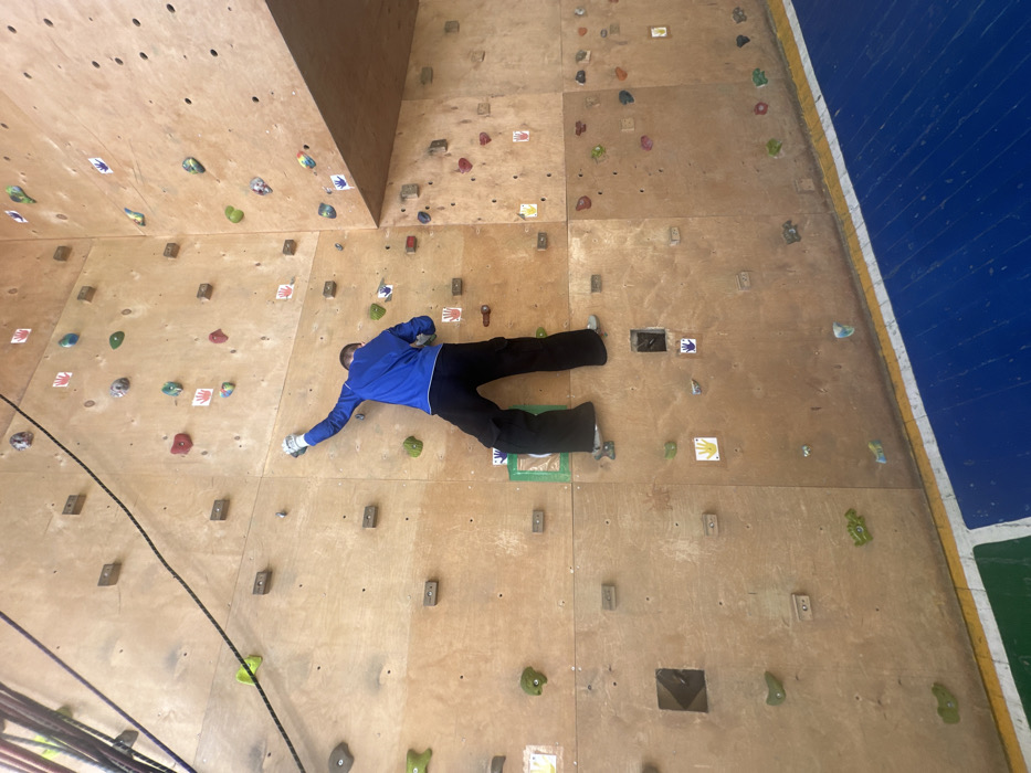 An individual is shown from behind, actively engaged in rock climbing on an indoor wall within a facility in Sarapul, Russia. The climber wears a blue long-sleeved shirt, black trousers, and light-colored footwear, with light-colored gloves on both hands. The right hand is extended upwards, gripping a grey-white climbing hold, while the left hand grasps a green hold. The right foot is positioned on a rectangular green hold, and the left foot rests on a smaller, light-colored hold. The climbing wall is constructed from light-brown wooden panels, featuring numerous climbing holds of various shapes and colors (red, green, blue, grey, orange, yellow). Small square stickers with colorful handprint symbols are affixed to the wall near certain holds. A black climbing rope is visible in the upper left section of the wall. The floor beneath the wall is covered with dark blue and green matting, separated by a yellow line.