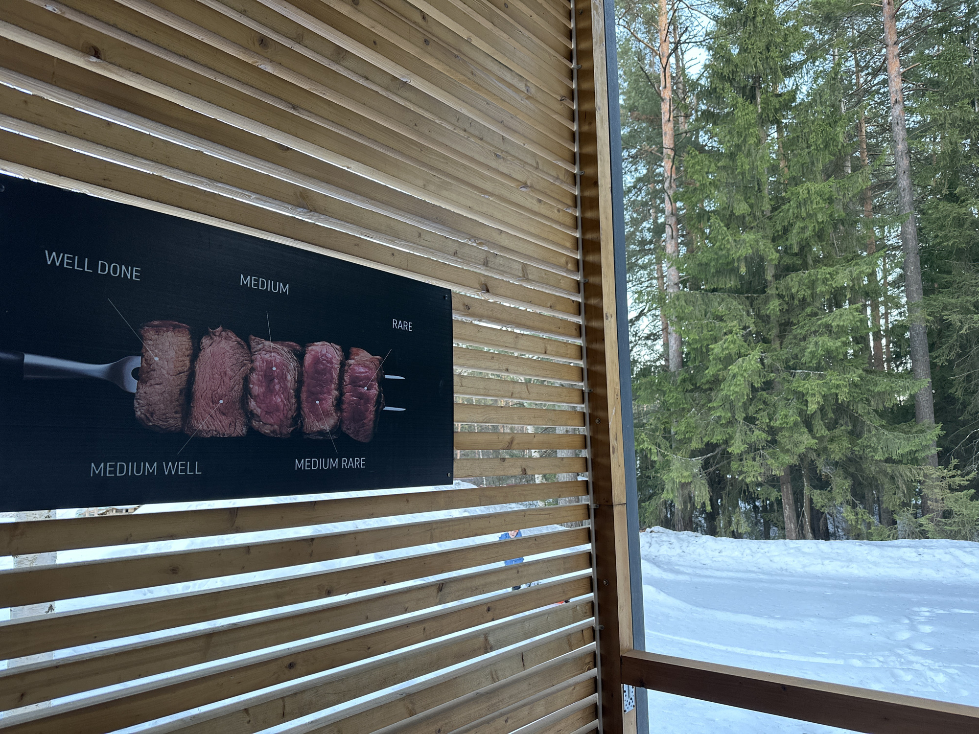 The image depicts a winter outdoor scene viewed from within a light-colored slatted wooden structure, likely in Прикамский, Russia. On the left, a vertical wooden slat wall features a dark rectangular sign displaying five steak cuts, arranged from "WELL DONE" to "RARE" with corresponding labels, and a partial image of a two-pronged fork. Through the horizontal wooden slats and an open section on the right, a snow-covered landscape is visible. A dense forest of coniferous trees occupies the background under a light blue sky. A small figure in a blue jacket is partially visible outdoors through the lower slats, situated on the snow. The immediate foreground on the right side includes a vertical wooden beam and a horizontal wooden railing, forming part of the structure.