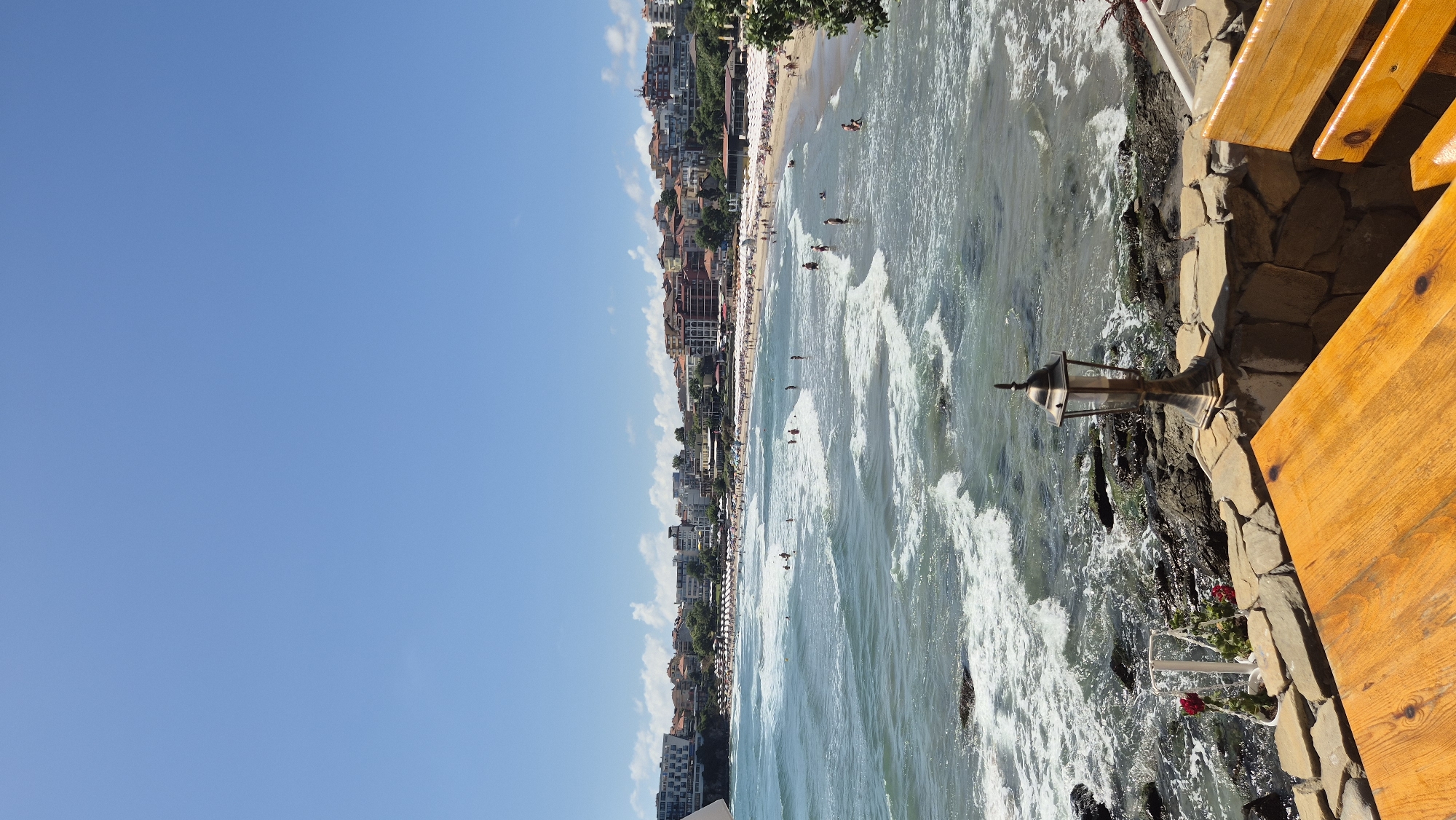 A bright, sunny day at a crowded beach in Sozopol, Bulgaria, is depicted from a slightly elevated vantage point. The foreground features a portion of a wooden table and a stone wall with an ornamental lamp. Beyond this, the ocean stretches out, with moderate waves breaking near the shore and white foam visible. Numerous people are scattered across the sandy beach and in the shallow water, with rows of white umbrellas providing shade along the shoreline. A cityscape of buildings with terracotta roofs and modern structures lines the coast. The sky is a clear, vibrant blue with a few wispy clouds.