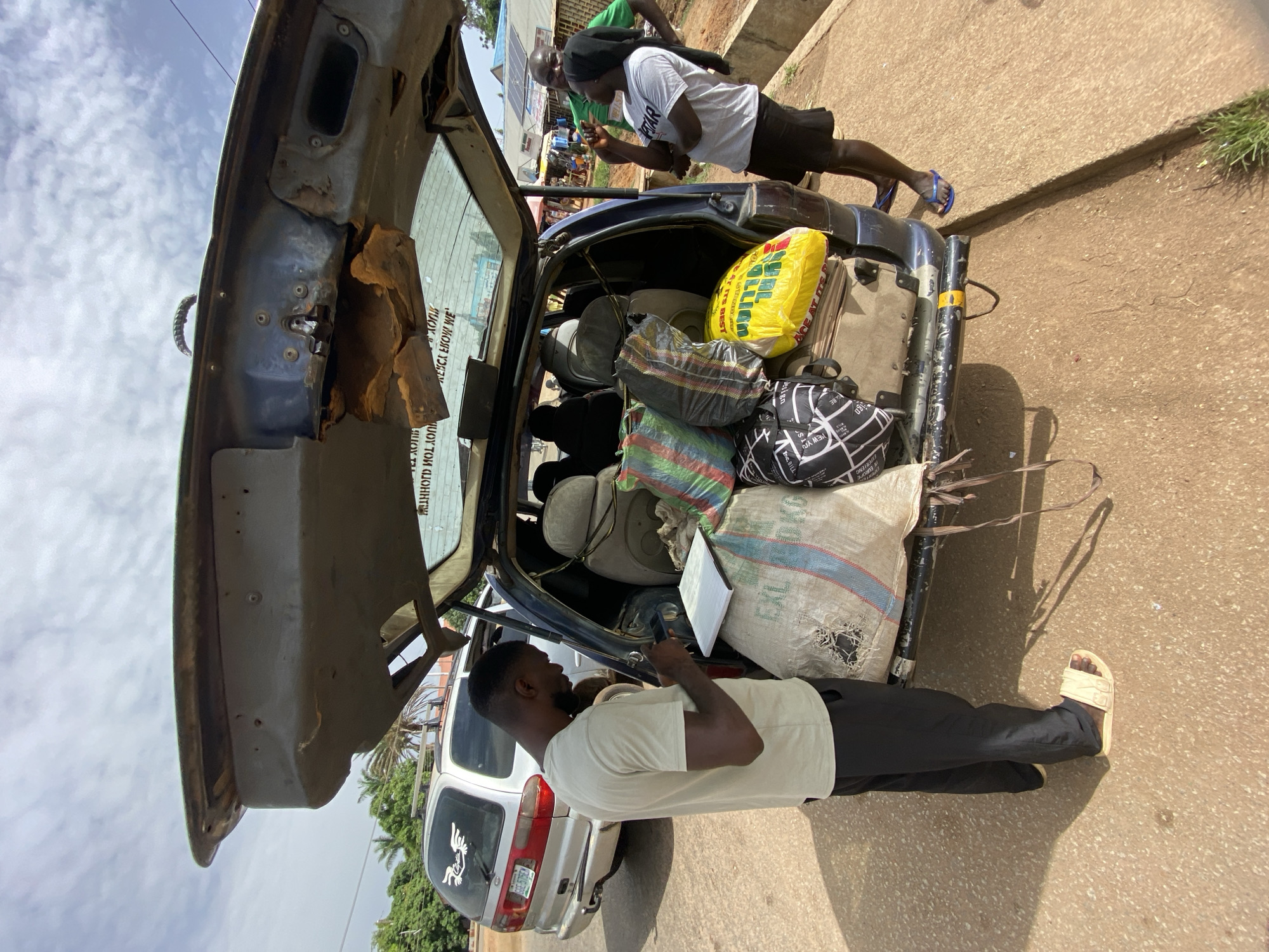The image shows a vehicle with an open trunk overloaded with various items, including bags and what appear to be seats, parked on a street. One person is standing near the back of the vehicle, looking at a phone. Another person is standing near the front of the vehicle. The setting appears to be outdoors on a sunny day. Text on the yellow bag: "BCE AT ITS BEST" "NCE AT DES". Text on the bag on the right side of the trunk: "EXL". The image text "WEBCA ESOW WE" appears to be a reflection from a window. Text on the black and white bag: "NEW YO". The license plate of the second vehicle is not readable. Based on the visible characteristics and context, the location is Bukuru, Nigeria.