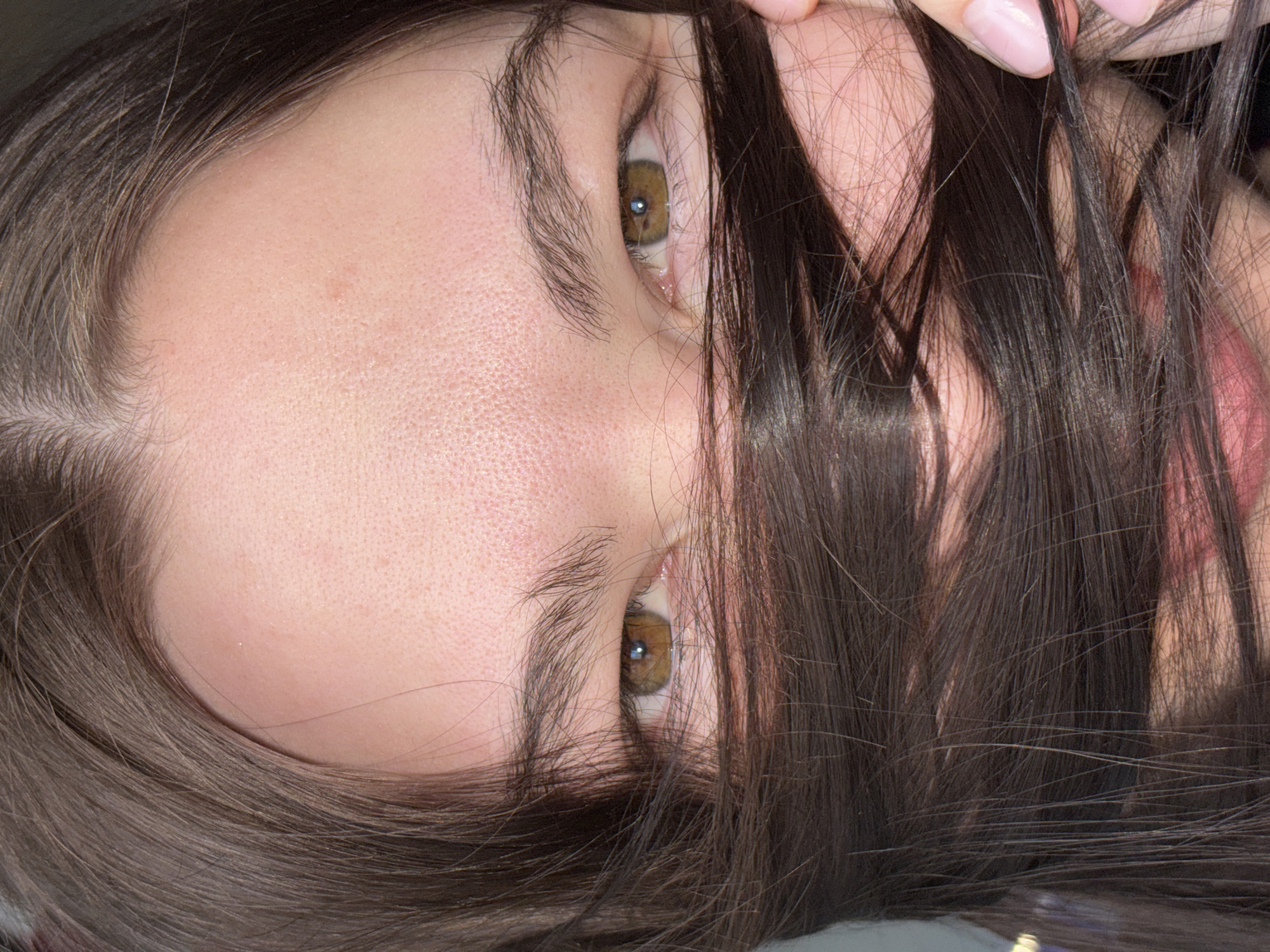 The image shows a close-up of a person's face, with dark brown hair partially obscuring it. The face is positioned at an angle, with the forehead and eyes visible. The eyes are hazel. A hand is holding some strands of hair near the top right of the image. No specific location in Perm, Russia, or any city is identifiable from the image.
