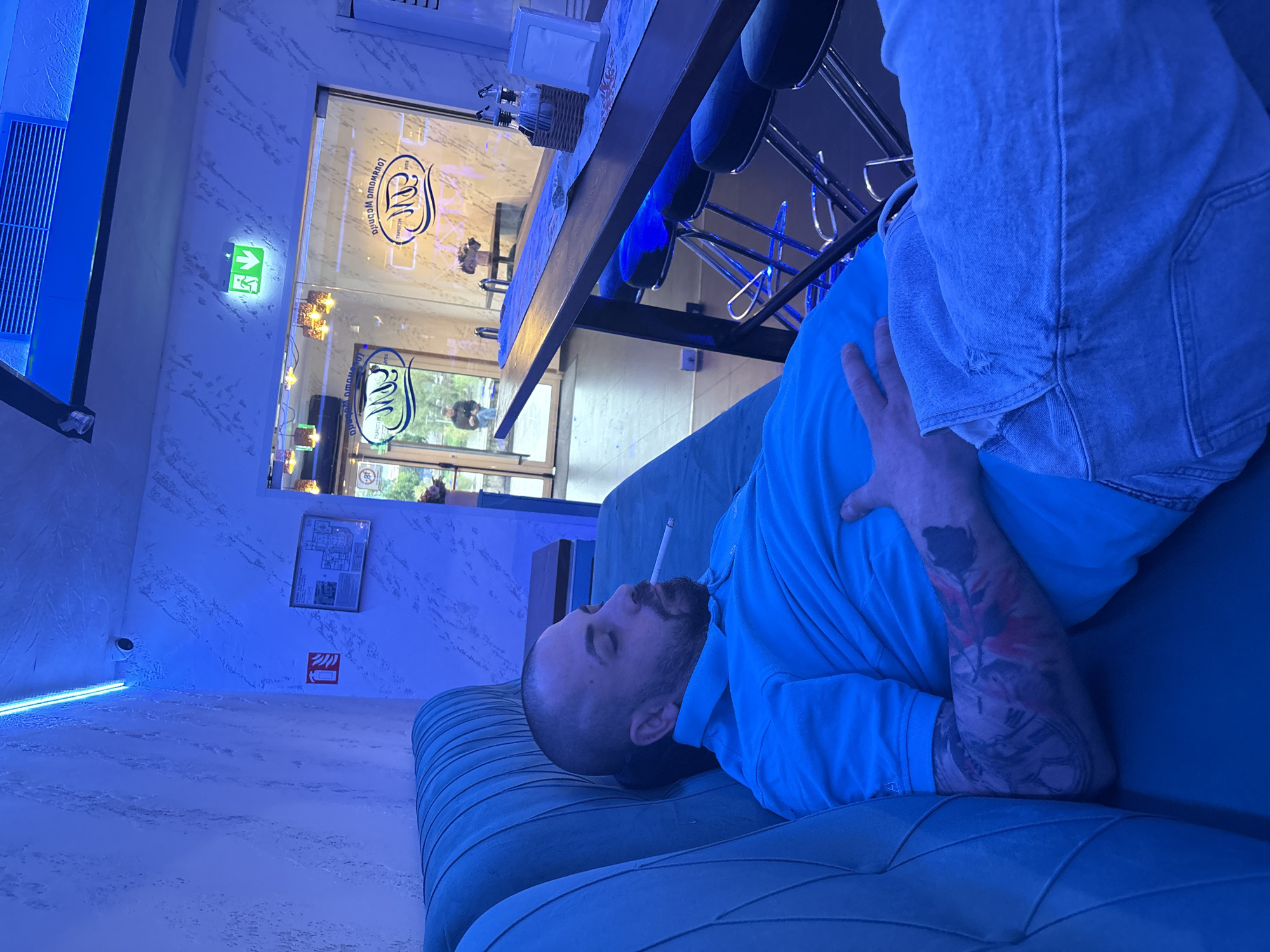 A man with a shaved head and beard lies on his back on a teal-colored sofa in a lounge or cafe. He is wearing a light blue polo shirt and light blue denim shorts, with visible tattoos on his left arm, including a rose and what appears to be a clock face. A lit cigarette is held between his lips. His right hand rests on his torso.
The interior space is bathed in a strong blue ambient light, likely from artificial sources like a neon strip visible along the left wall. The walls have a textured, marbled appearance in white and light blue tones. In the background, a long wooden bar counter with several high stools is visible. Behind the bar is a large glass door or partition through which daylight and the blurred figure of another person can be seen outside. The glass features a stylized logo that resembles an "M" or "W" and text, including the word "MELODIA" (ΜελΩΔΙΑ) and "LOU MAILIA" (ΛOUMAIΛIA), appearing both mirrored and right-reading.
On the wall to the left, a green emergency exit sign with an arrow pointing right is illuminated. Further down, a small red fire extinguisher symbol is present, accompanied by an unreadable text sign. A framed document or map hangs on the wall near the top right. A small basket with napkins or condiments sits on the bar counter. The man appears relaxed or contemplative in the cool-toned environment.
