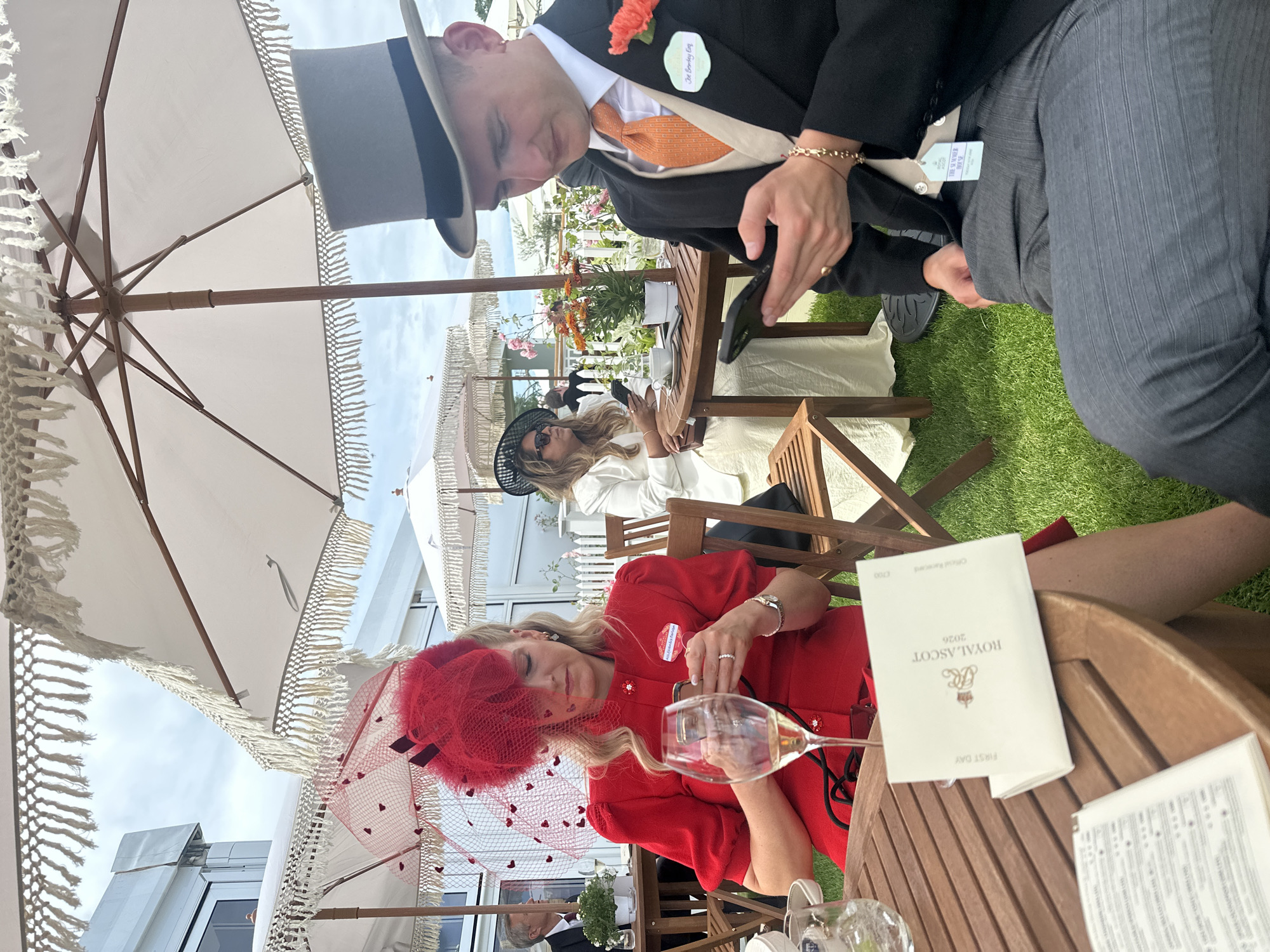 The image depicts a scene at Royal Ascot, a prestigious horse racing event in the United Kingdom. Three people are visible, two women and one man. The primary focus is on a woman in the foreground wearing a striking red dress and a matching elaborate red fascinator. She is looking down at her phone, with a glass of champagne beside her. A menu, identifiable as "ROYAL ASCOT 2026 Official Racecard FIRST DAY", rests on the wooden table in front of her.
To her left, a woman in a white outfit and a black hat is also seated at a table, partially obscured. To the right and slightly behind, a man in a formal suit, complete with a top hat and an orange tie, is also looking down, possibly at his phone as well. His attire suggests he is participating in the event's traditional dress code.
The setting appears to be an outdoor hospitality area, characterized by wooden tables and chairs, and large white parasols with fringed edges providing shade. The grass area suggests a well-maintained outdoor venue. The weather seems pleasant, with bright daylight visible through the umbrellas, indicating it is likely daytime. The overall atmosphere suggests a social and celebratory occasion, typical of high-profile sporting events where attendees often dress formally. The presence of champagne glasses and a racecard further reinforces the context of the event.