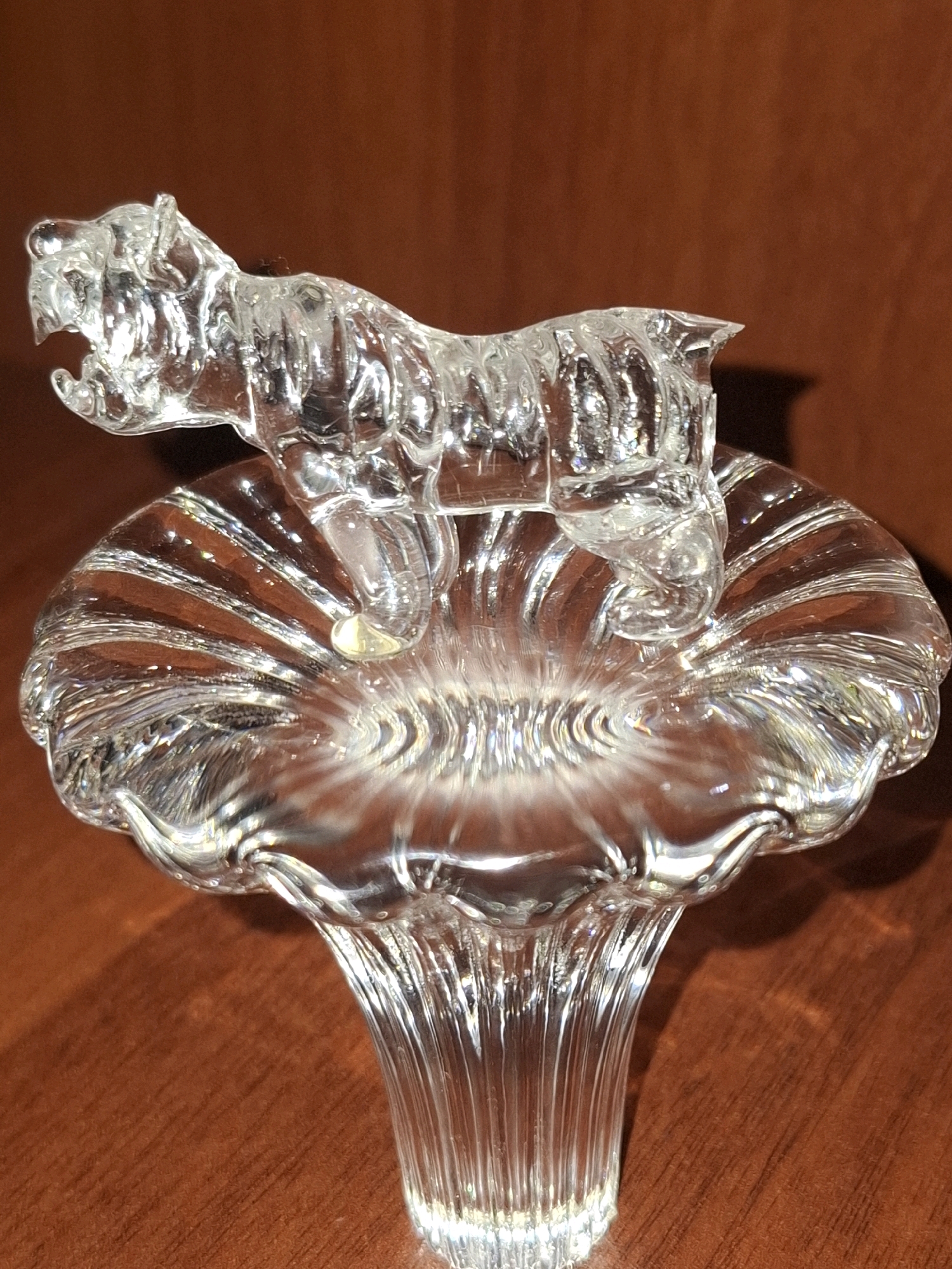 A clear glass figurine of a tiger stands on top of a clear glass mushroom-shaped pedestal. The tiger is sculpted in a dynamic pose, with its mouth open and its body rippling with detail. The pedestal has a fluted design on its cap and a ribbed stem, creating a play of light and shadow. The background is a warm, dark brown, likely a wooden surface, which contrasts with the transparency of the glass objects. There are no people or any other objects visible in the scene. The image appears to be a product shot or a still life of decorative items. The location in Almaty, Kazakhstan, is not discernible from the image.