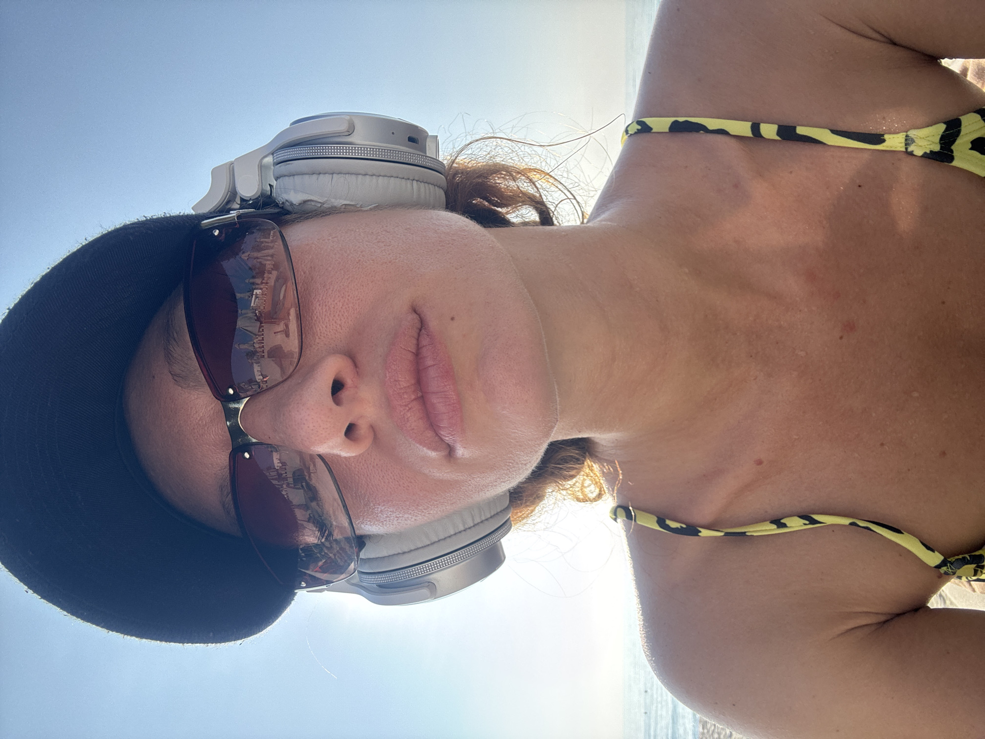 A woman is posing for a selfie on a sunny day in Baan Chaweng, Thailand. She is wearing a black baseball cap, sunglasses with reflective lenses, and headphones. A yellow and black bikini top is visible on her shoulder. The ocean is in the background.