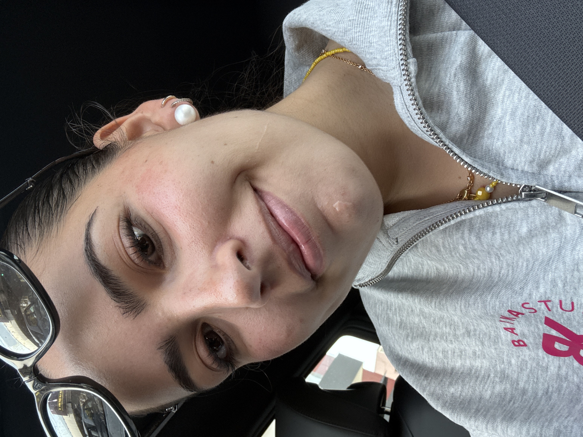 The image shows a woman indoors, possibly in a car, wearing glasses, a grey hooded jacket, and jewelry (earring and necklace). The text on the jacket reads "BANASTUA". The location is possibly Puebla, Mexico, based on the brand.