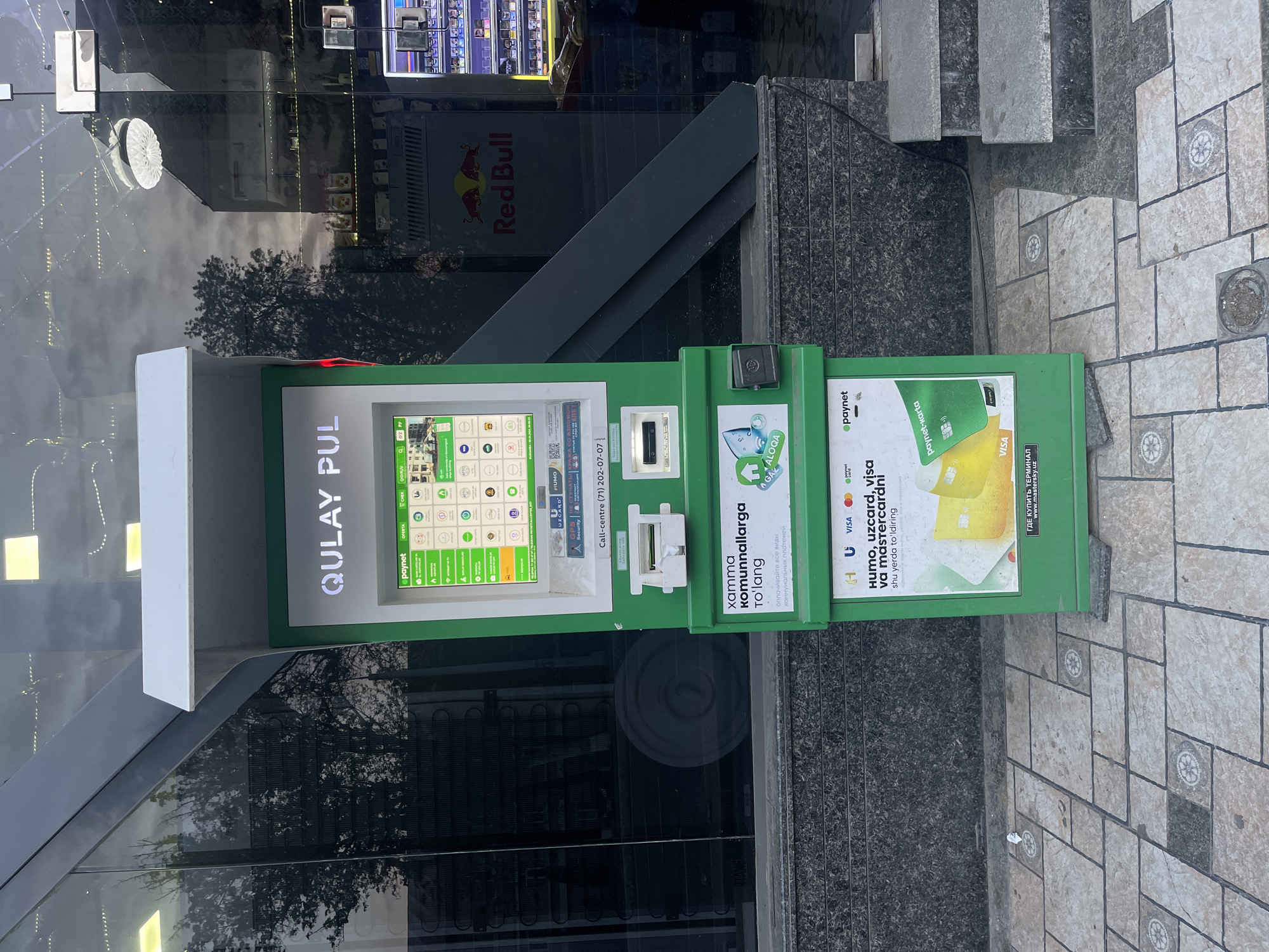The image shows a green "Qulay Pul" payment kiosk, a common sight in Tashkent, Uzbekistan. The kiosk features a touchscreen interface with icons representing various payment options. Adjacent to the screen is a card reader and a slot for bills. Advertisements promoting electronic payments and card usage, including HUMO, UZCARD, Visa, and Mastercard, are displayed on the side of the kiosk in Uzbek and Russian. The kiosk is located on a city street with a tiled sidewalk and granite stairs leading to a building with a storefront display, including a Red Bull advertisement.