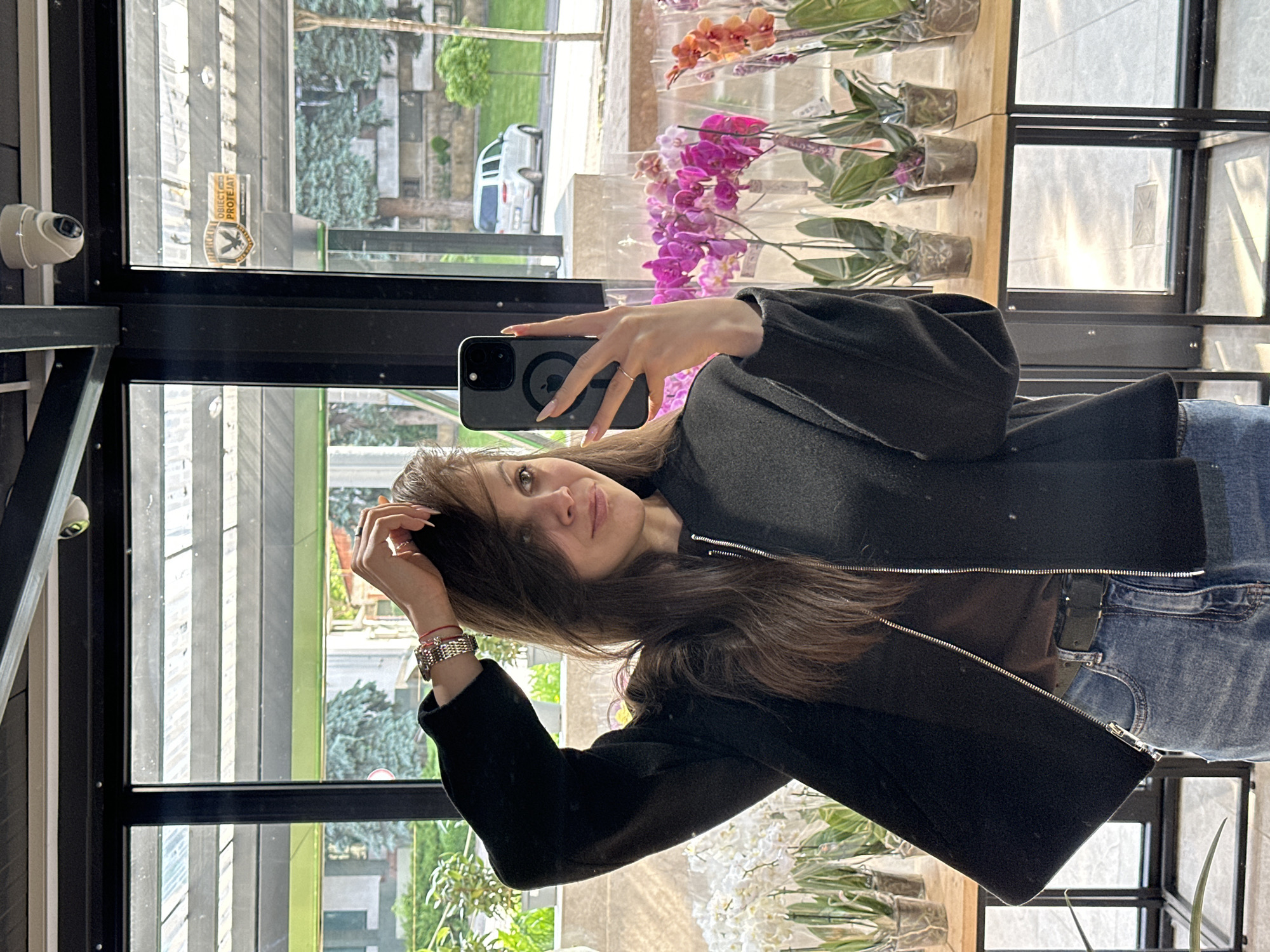 The image depicts a mirror selfie taken by a female subject within what appears to be a floral shop or greenhouse in Chișinău, Moldova.
The subject is a female with long, dark brown hair, some strands falling across her forehead. She is wearing a black zippered jacket over a brown top, paired with blue jeans and a dark belt. On her left wrist, she wears a metallic-banded wristwatch and a red string bracelet. Her fingernails are manicured with a light-colored polish. She holds a dark-colored smartphone, consistent with an iPhone model (showing a MagSafe ring reflection), in her right hand, positioned to capture the reflection. Her left hand is raised, with fingers gently touching her hair on the right side of her head. She exhibits a subtle smile, looking directly at the phone's camera reflection.
The interior environment features large glass windows, through which an outdoor scene is reflected. Inside, numerous orchids are displayed in clear pots on light-colored wooden shelves with black metal frames. White orchids are visible on the viewer's left, while magenta, purple, and orange orchids are on the viewer's right. The ceiling structure is dark, with two white dome-shaped security cameras mounted at the top. A rectangular sticker is affixed to the glass window, bearing a shield logo with an eagle and the text "OBIECT PROTEJAT" (Protected Object), written in Romanian, indicating a location in Moldova.
The reflected exterior scene includes a paved road, a white van parked on the street, and various green trees and bushes, along with distant buildings, under natural daylight.