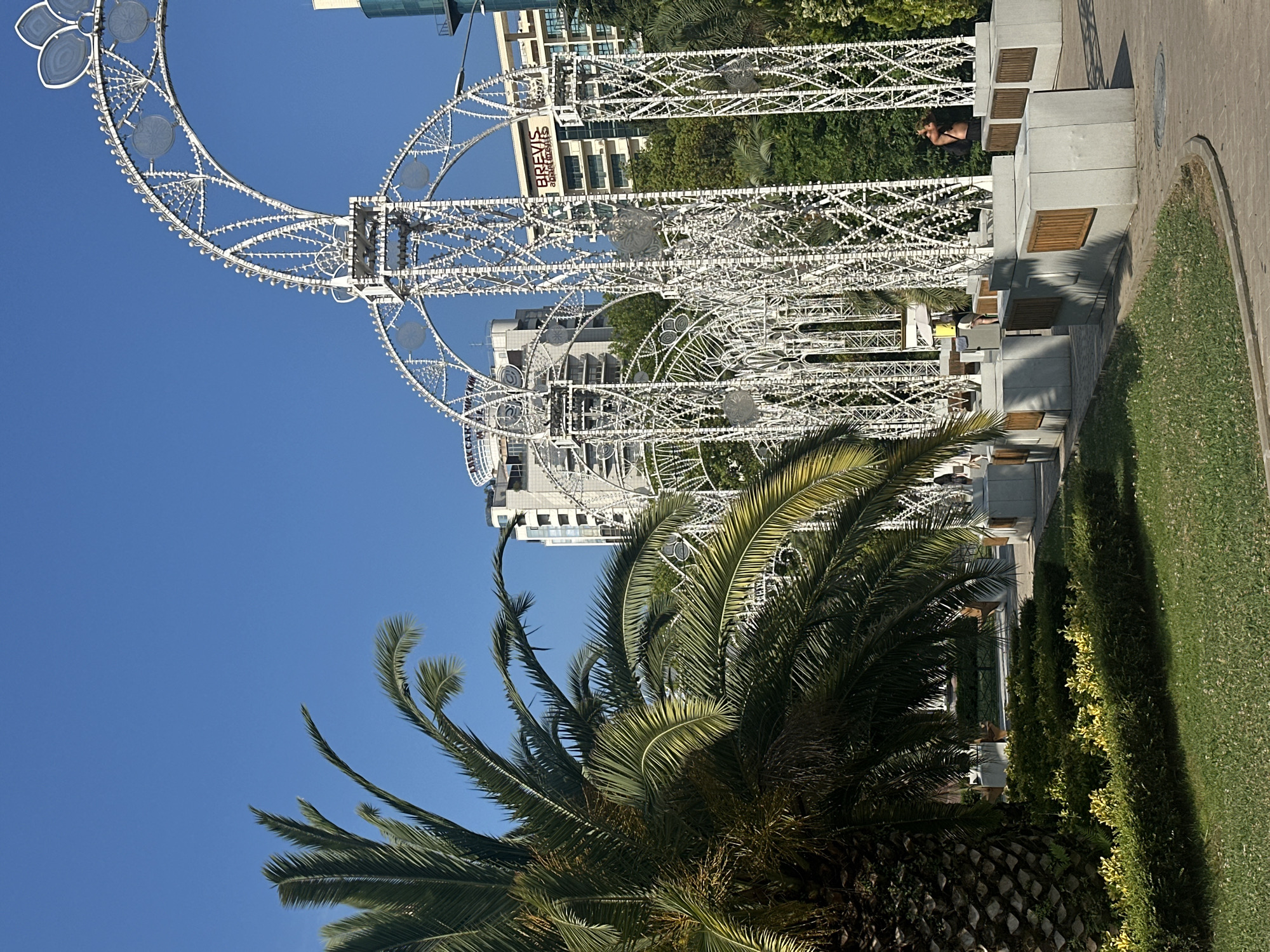 In the city of Sochi, Russia, a palm tree stands to the left of a large white ornate archway structure. The archway is illuminated with many small lights and is decorated with circular patterned elements. Behind the archway, modern buildings are visible, one of which displays the text "BREVIS apartments." Green foliage and neatly trimmed bushes are present in the foreground. A pathway with concrete benches, some featuring wooden accents, is situated around the archway structure. A person, partially obscured, is visible on the right side of the frame, near one of the benches. The sky is clear and blue.