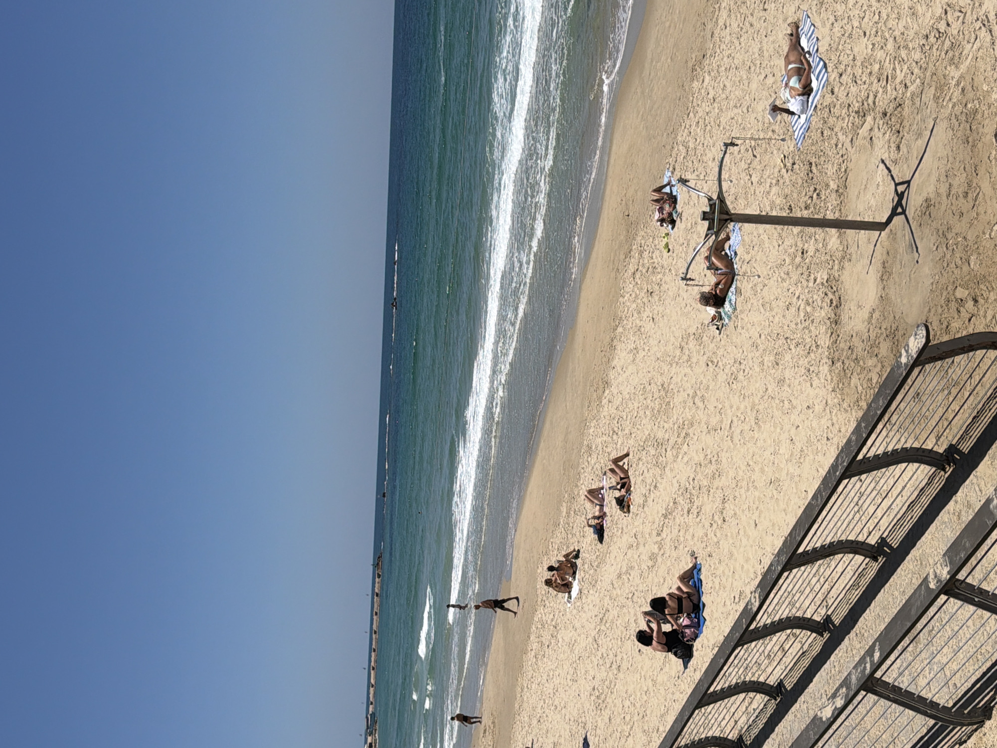The image depicts a sunny day at a beach in Tel Aviv, Israel. The foreground shows a dark metal railing casting shadows on the sand. Several people are visible on the beach, scattered across the sand. In the middle ground, a group of individuals are sunbathing on towels, some lying down, others sitting. Further back, a man walks along the shoreline, and a few other people are present near the water's edge. The sea is blue with gentle waves, and in the distance, a breakwater structure extends into the water. The sky is a clear, bright blue.
