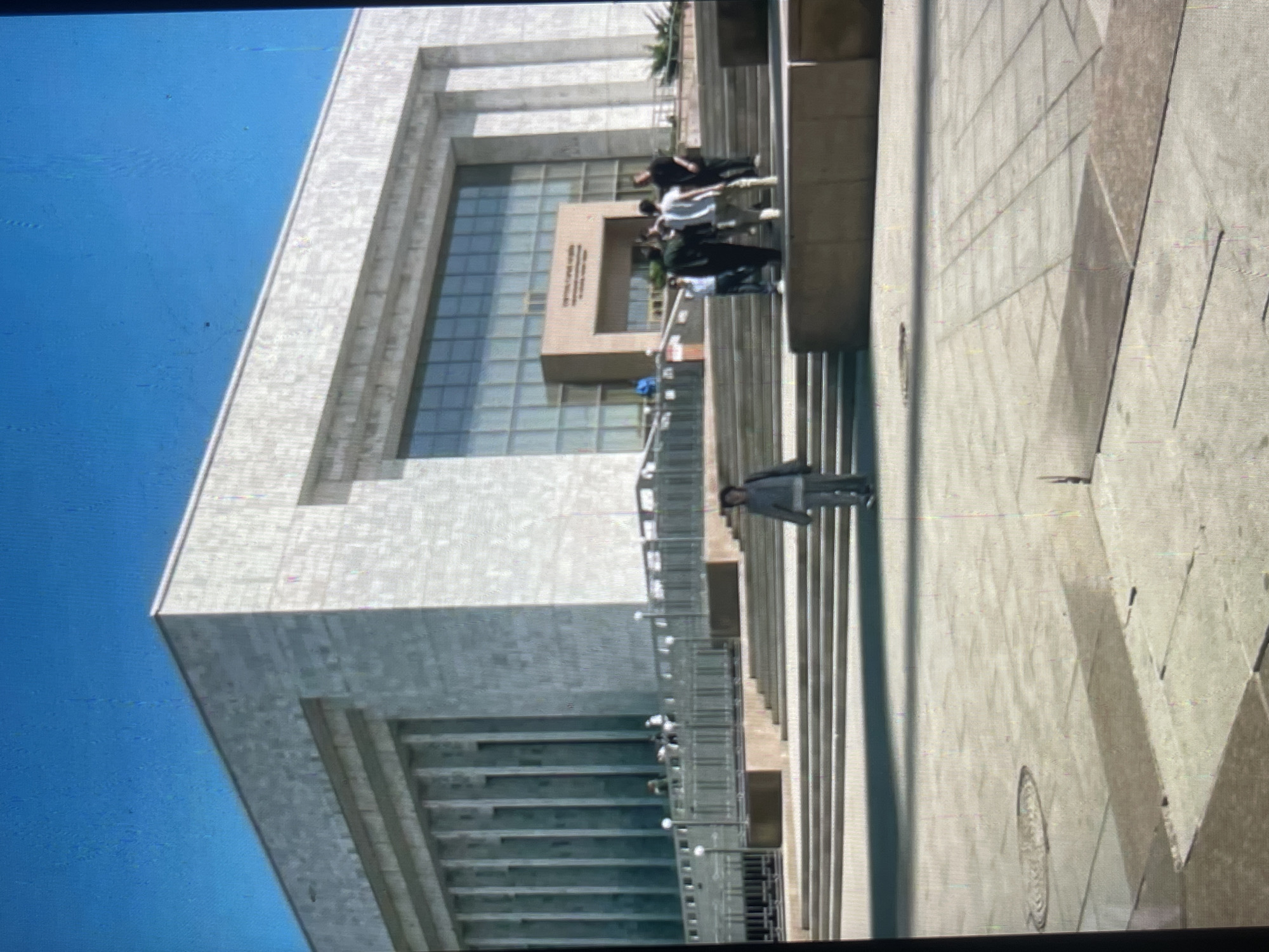 This image captures the exterior of the National Library of the Kyrgyz Republic in Bishkek, Kyrgyzstan. The building features a large, modern facade clad in light-colored stone, with a prominent central section containing a wide glass entrance. A wide, multi-tiered stone staircase leads up to the library's entrance from the plaza in front. Several individuals are visible on the steps and in the plaza. In the foreground, a person in a light-colored jacket and pants walks down the steps. Further up the stairs, a group of people are gathered, and some are entering the building. The sky above is a clear, bright blue. The plaza area in the foreground is paved with large square tiles and shows distinct shadows cast by the sun.