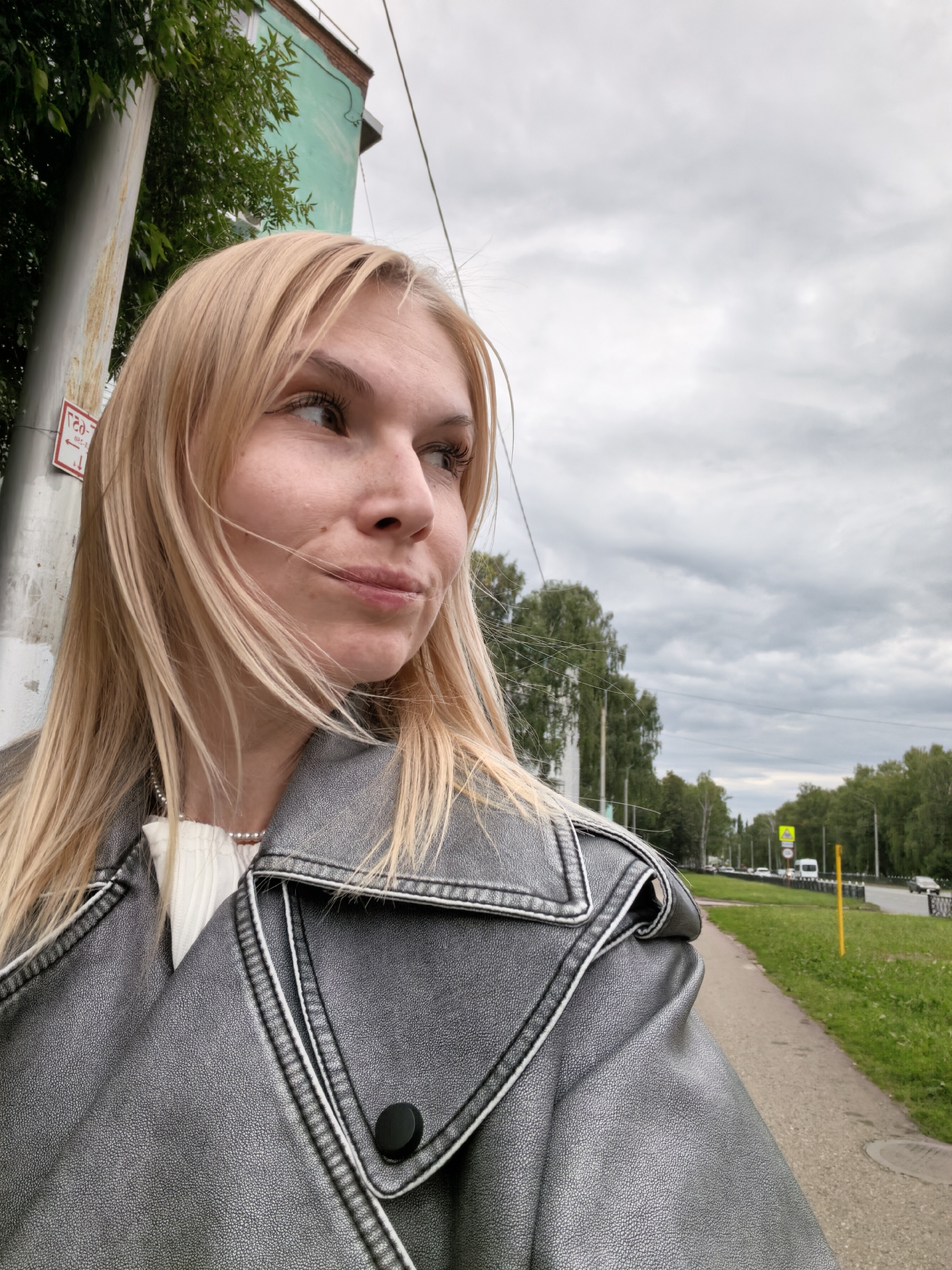 A young woman with blonde hair, wearing a grey leather jacket and a white top with a pearl necklace, poses for a selfie outdoors. She is looking to her right, with her head tilted slightly. The background shows a cloudy sky, trees, a sidewalk, and a road with cars. The image was taken on a street in Sterlitamak, Russia, likely near a building with a green facade. A sign on a pole in the foreground is partially visible, showing Cyrillic text.