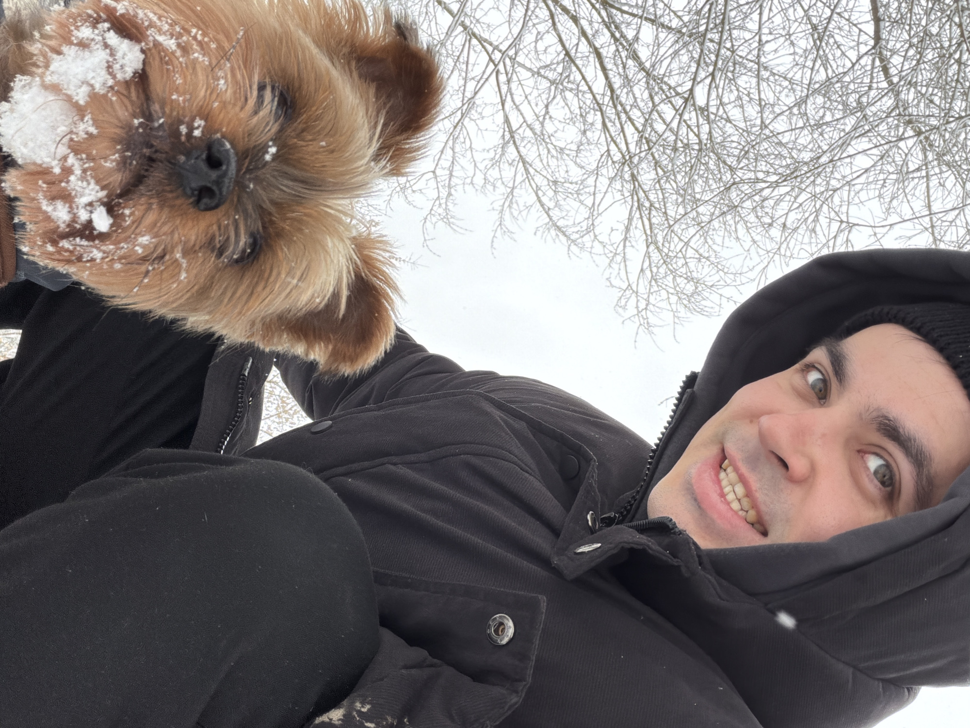 A selfie captures a man and his dog in a snowy outdoor setting. The man, wearing a black hooded jacket and a beanie, is smiling at the camera. His dog, a small breed with brown fur and snow on its muzzle, is positioned above him in the frame. The background reveals a snow-covered scene with bare tree branches against an overcast sky. While the precise location in Izhevsk, Russia is not discernible from the image, the presence of snow and bare trees indicates a winter setting.