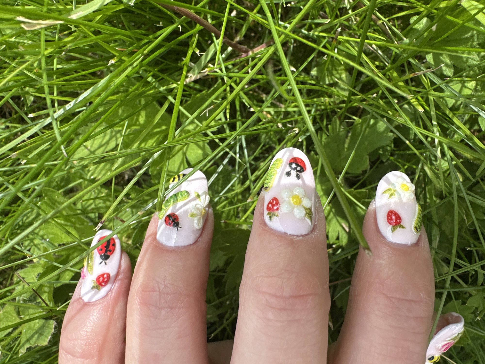 The image shows a close-up of a person's hand with fingernails decorated with a 3D nail art design featuring ladybugs, strawberries, and white flowers on a light pink base. The hand is positioned over green grass, suggesting an outdoor setting. The lighting indicates it is daytime, with the sun casting shadows. There is no discernible location in Moortown, Ireland, or any other specific urban setting visible in the image. The focus is on the detailed nail art and the natural, grassy environment.