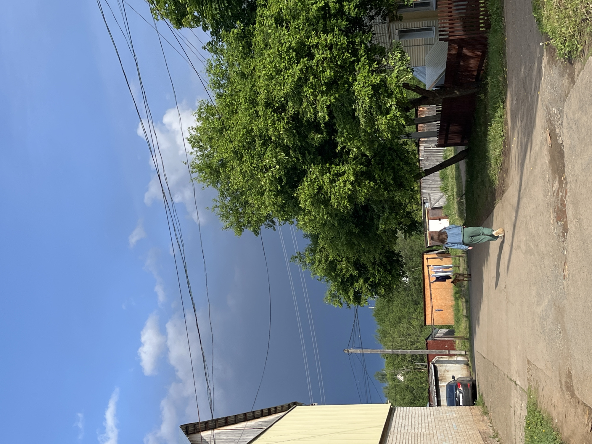 A street scene in Фоки, Russia is depicted in this rotated medium shot. A concrete path or road stretches through the frame with a woman, clad in a denim jacket and green pants, walking on the path. The sky transitions from a bright blue with light clouds to a dark, ominous grey, suggesting an approaching storm. Overhead power lines cut across the sky. Adjacent to the path are various buildings, including a light-colored, possibly yellow, structure with a slanted roof. Additional buildings and structures are visible further down the street, with a car parked next to one. A large green tree looms over the path and adjacent buildings. Clothes appear to be drying outside of the structure.