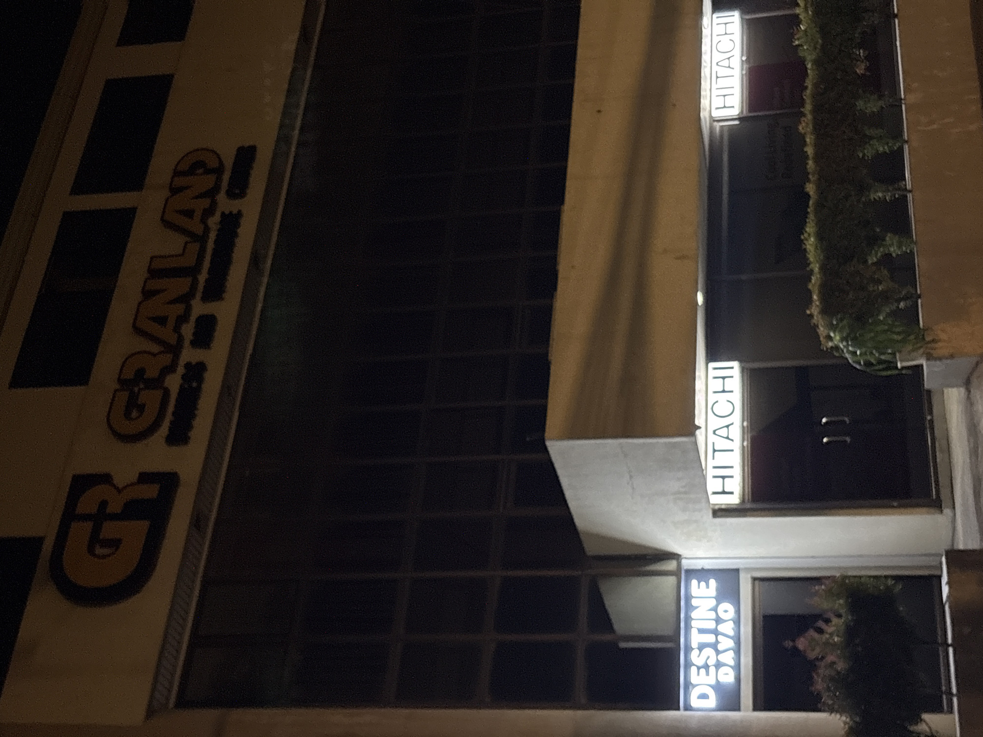 The image shows the exterior of the Granland Business and Warehouse Center in Davao City, Philippines, at night. The building has a large, illuminated sign at the top with the letters "GR" followed by "GRANLAND." Below this, smaller text reads "BUSINESS AND WAREHOUSE CENTER." On the lower part of the building, there are two illuminated signs for "DESTINE DAVAO" on the left and "HITACHI" on the right. The facade features large, dark windows. In front of the "DESTINE DAVAO" entrance, there is some greenery. The "HITACHI" entrance appears to be a set of double doors. The scene is lit by artificial lighting, creating a stark contrast between the illuminated signs and the dark surroundings.