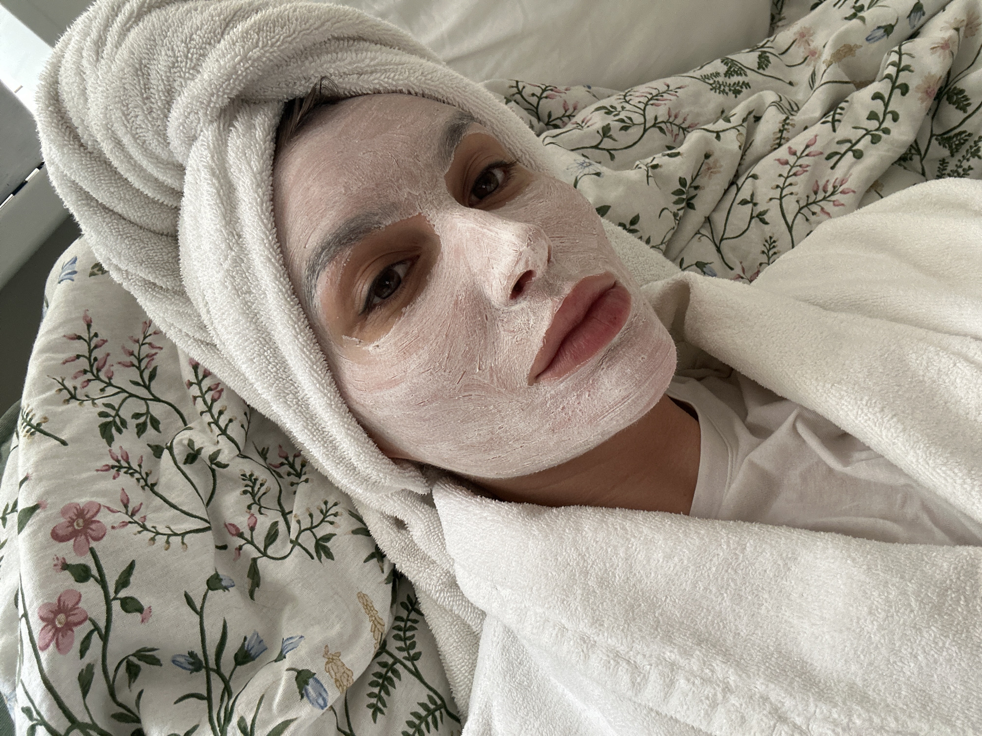 A woman is lying on a bed covered in a floral-patterned sheet, wearing a white facial mask and a white towel wrapped around her head. She is wearing a white robe or shirt. The location is not identifiable as Moscow, Russia.