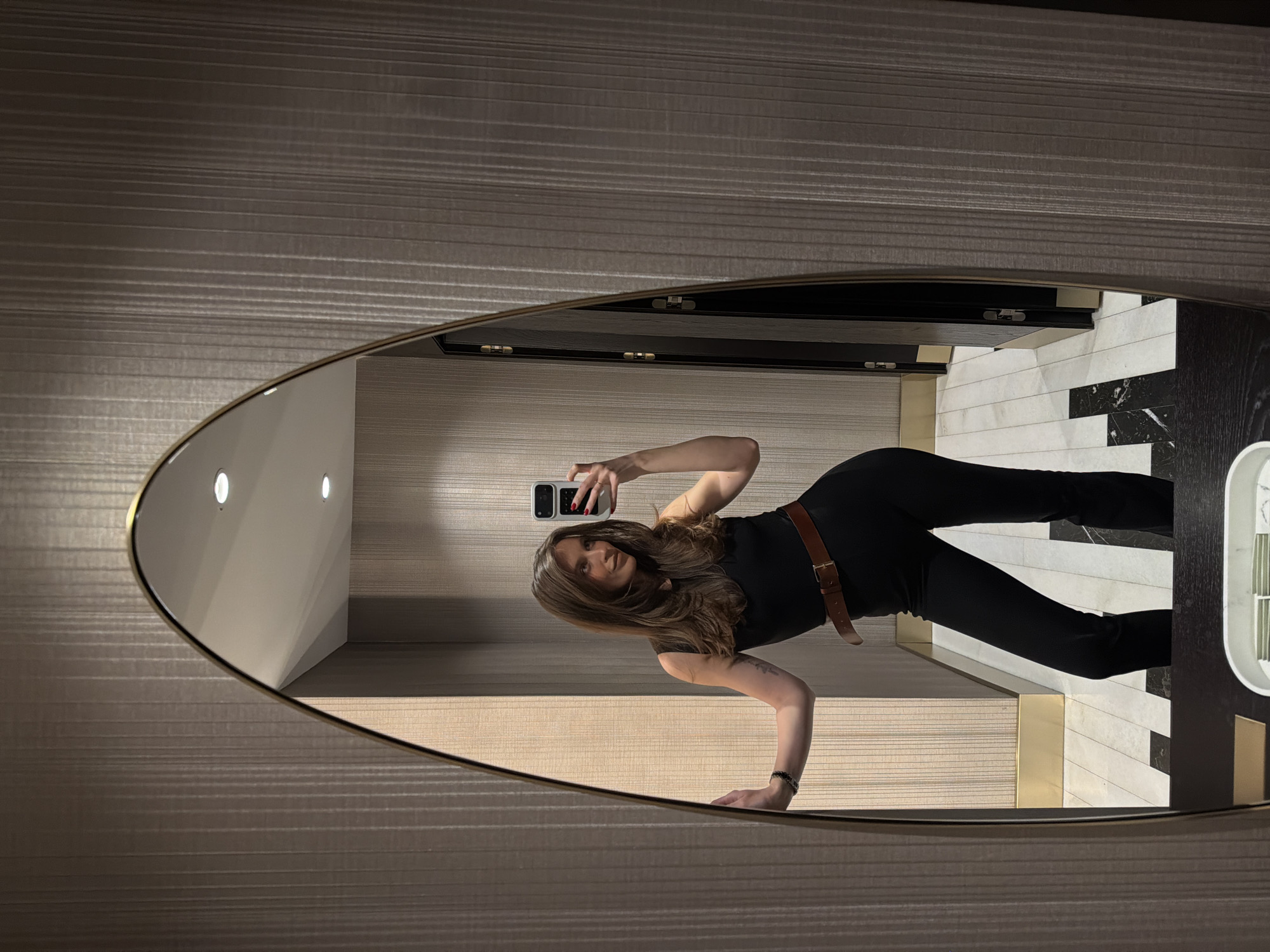 A female subject stands in a well-lit indoor space, captured via a mirror selfie. The subject has long, dark brown hair and is dressed in a black sleeveless top, black pants, and a brown leather belt. Her right hand holds a smartphone with a white case and a black rectangular accessory on its rear; red nail polish is visible on her fingers. Her left arm is extended, with her hand resting against a wall to the left of the mirror. A dark, beaded bracelet is present on her left wrist.
The primary object is a full-length, elongated oval mirror with a gold-toned frame, reflecting the subject and the immediate environment. The walls are covered in a textured, vertical striped wallpaper in a muted taupe or warm gray tone. The floor features a black and white geometric pattern, consisting of black rectangular segments on a white background, indicative of marble or high-end tile. A gold-toned metallic baseboard runs along the bottom of the walls. The ceiling is white and includes recessed circular light fixtures. In the reflection behind the subject, several dark-colored doors with visible gold hinges are present, suggesting a multi-stall restroom or a corridor with multiple entrances. A white container, possibly a tissue dispenser, is partially visible in the foreground at the base of the mirror.
This scene is located within the **Dukley Hotel & Resort in Budva, Montenegro.**
