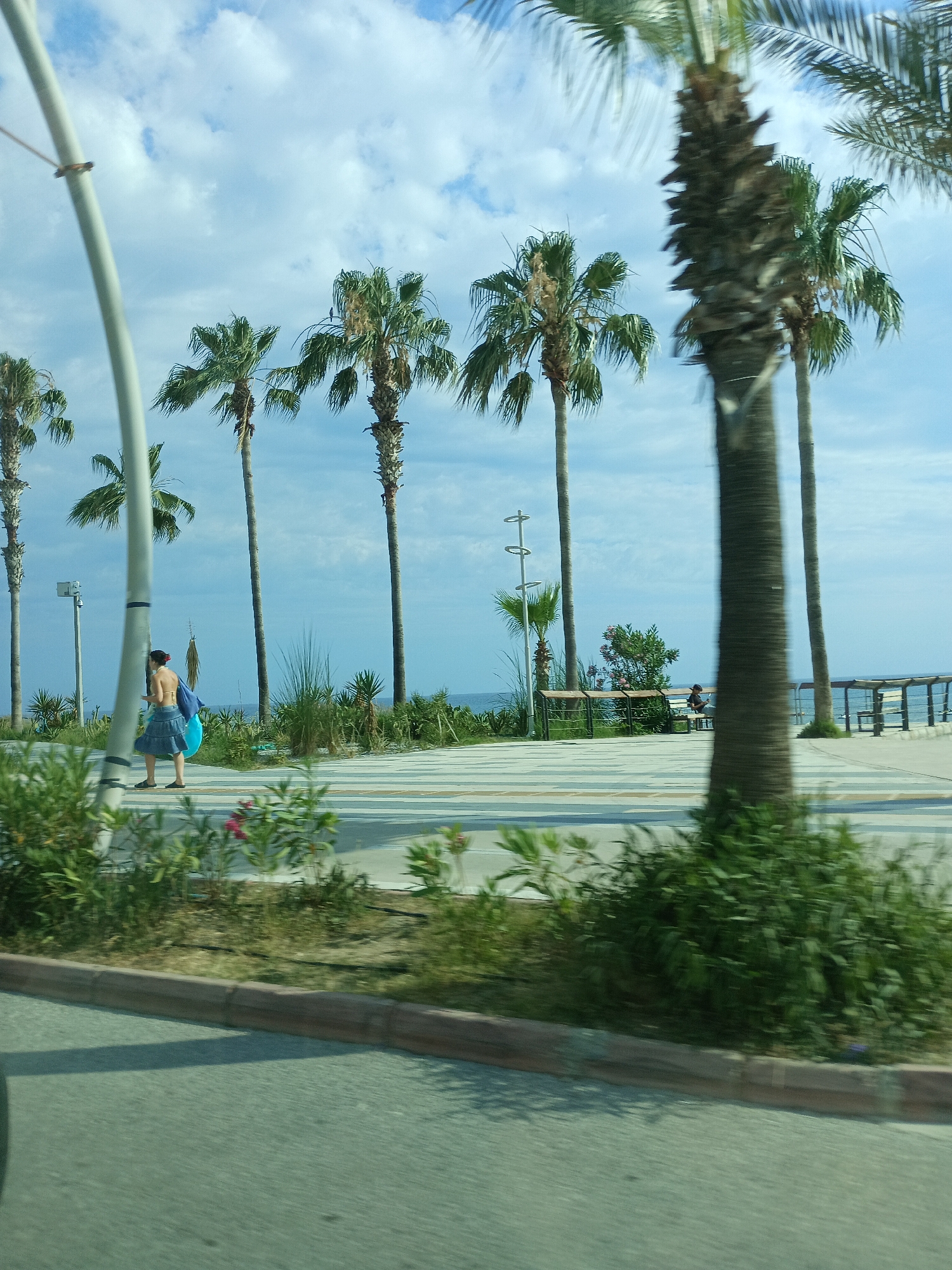 This image captures a sunny day in Alanya, Turkey, likely in the late afternoon given the warm light and long shadows. The scene depicts a pedestrian walkway with palm trees lining a beach or coastal area, with the blue sea visible in the background.
In the foreground, a person is walking along the path, carrying a bright blue object, possibly a beach mat or towel. They are wearing a bikini top and a denim skirt. Further down the walkway, another person is seated on a bench, appearing to be relaxed. The walkway itself is paved with light-colored tiles.
The weather is clear with scattered clouds in a bright blue sky. The palm trees, characteristic of a tropical or Mediterranean climate, are a dominant feature of the landscape. The presence of benches and a railing overlooking the sea suggests a public promenade or park area. The overall atmosphere is one of leisure and vacation, with the natural beauty of the coast providing a serene backdrop. There is no visible text in the image.