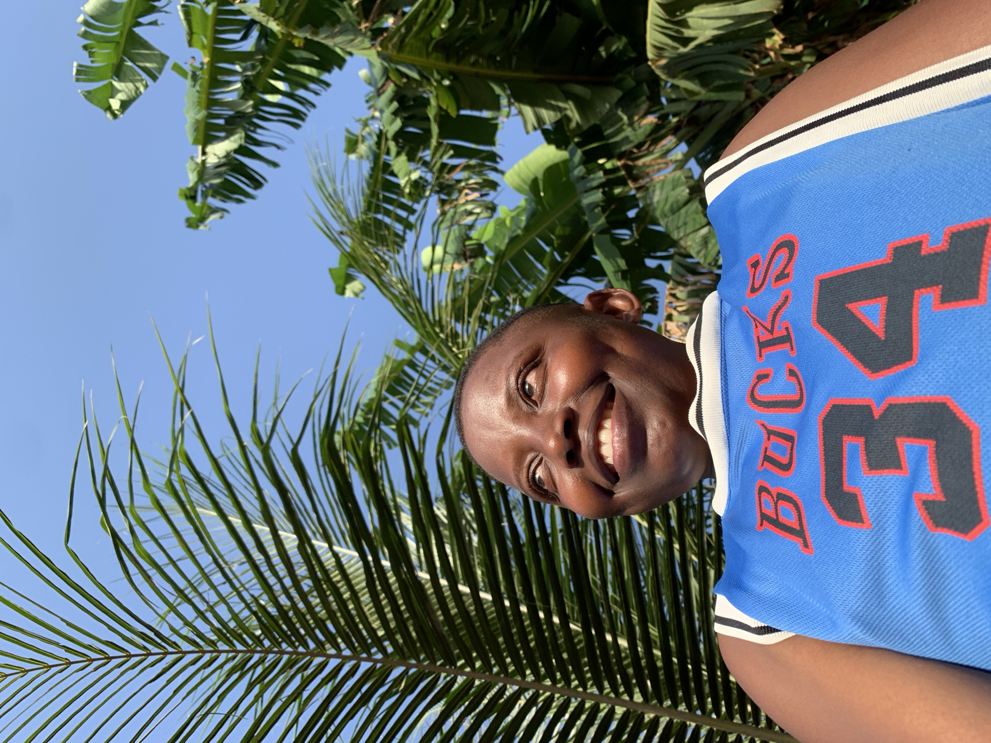 A person wearing a blue basketball jersey with the word "Bucks" and the number "34" on it is smiling at the camera. The person is positioned in front of a backdrop of palm trees and a clear blue sky. The photo appears to be taken from a low angle, looking up towards the person. There is no verifiable evidence to confirm the location is Ikorodu, Nigeria.