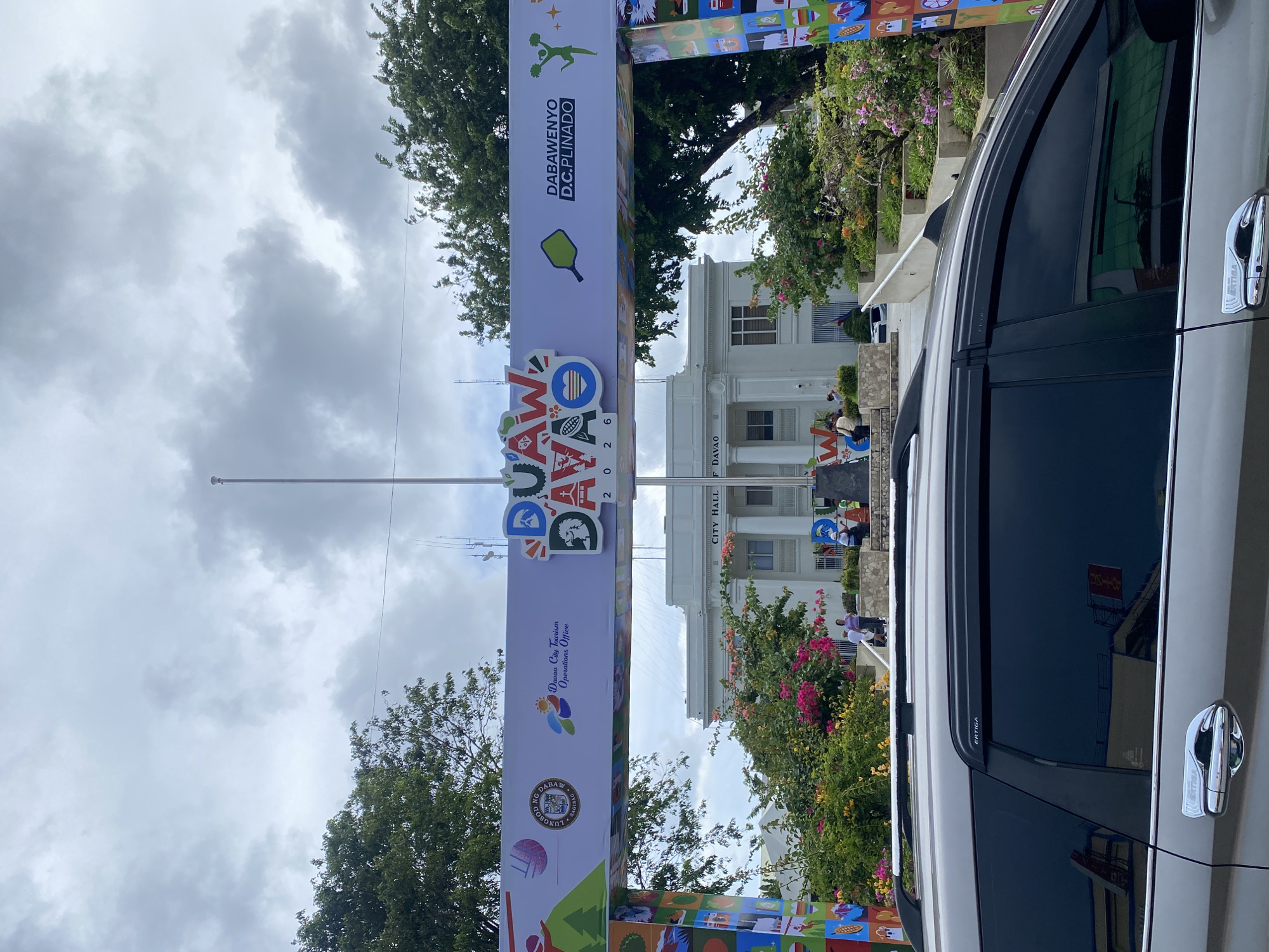 The image shows the City Hall of Davao City, Philippines, with a large banner overhead. The banner features the words "DUAW DAVAO" and "DABAWENYO D.C.PLINADO", suggesting an event or celebration. The City Hall is a white, two-story building with a colonnaded entrance. In front of the building, there are stairs, a planter with flowering bushes, and some people. A silver SUV is parked in the foreground, obscuring the lower part of the scene. The sky is cloudy.