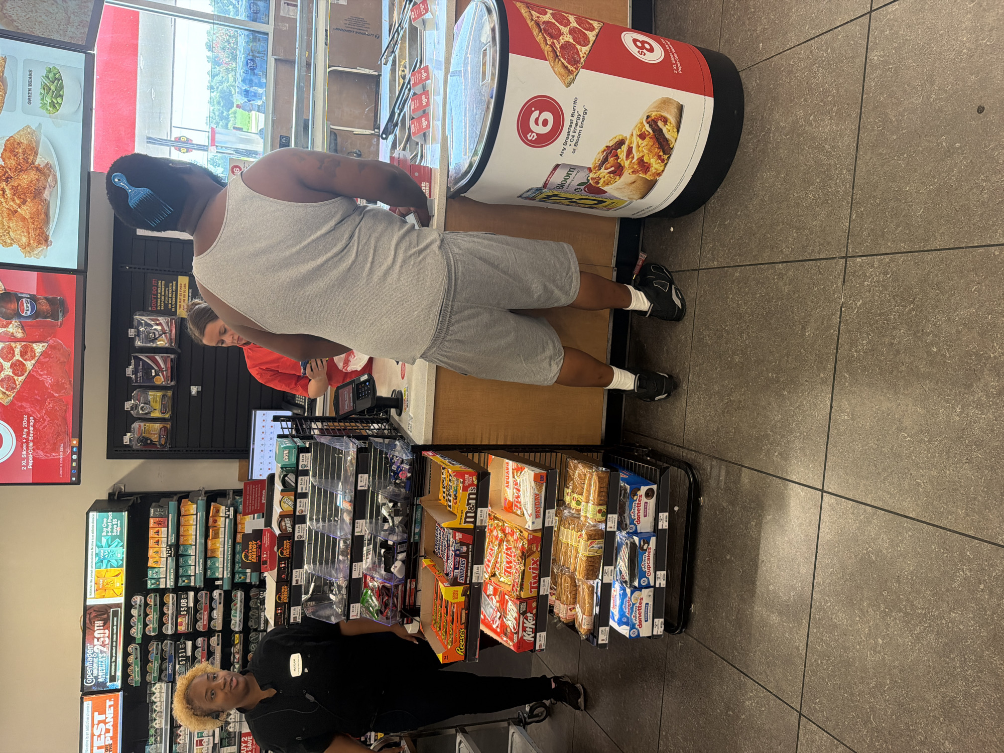 The image depicts the interior of a convenience store, likely a 7-Eleven, located in Hayti, Missouri, United States of America. A female employee, wearing a black uniform with a name tag reading "MAXIAM," stands behind a counter, interacting with a customer. The customer, a male wearing a grey tank top and grey shorts, is facing away from the camera, his back to the viewer. He has a blue hair pick in his hair. Various merchandise, including candy bars, packaged snacks, and drinks, are displayed on shelves and in refrigerated units throughout the store. A large promotional graphic for breakfast burritos and pizza is visible on a cylindrical display near the customer. The store appears to be well-lit and stocked.