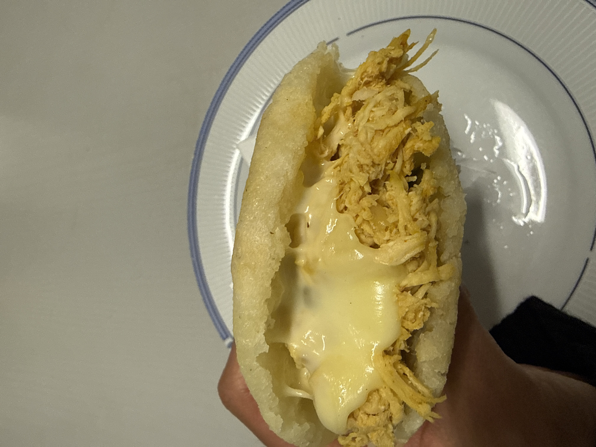 The image shows a hand holding an arepa filled with shredded chicken and cheese. The arepa is partially opened to reveal the filling. A white plate with a blue rim is visible in the background. The background also includes a plain white surface. There is no information to indicate the location is Barcelona, Spain.
