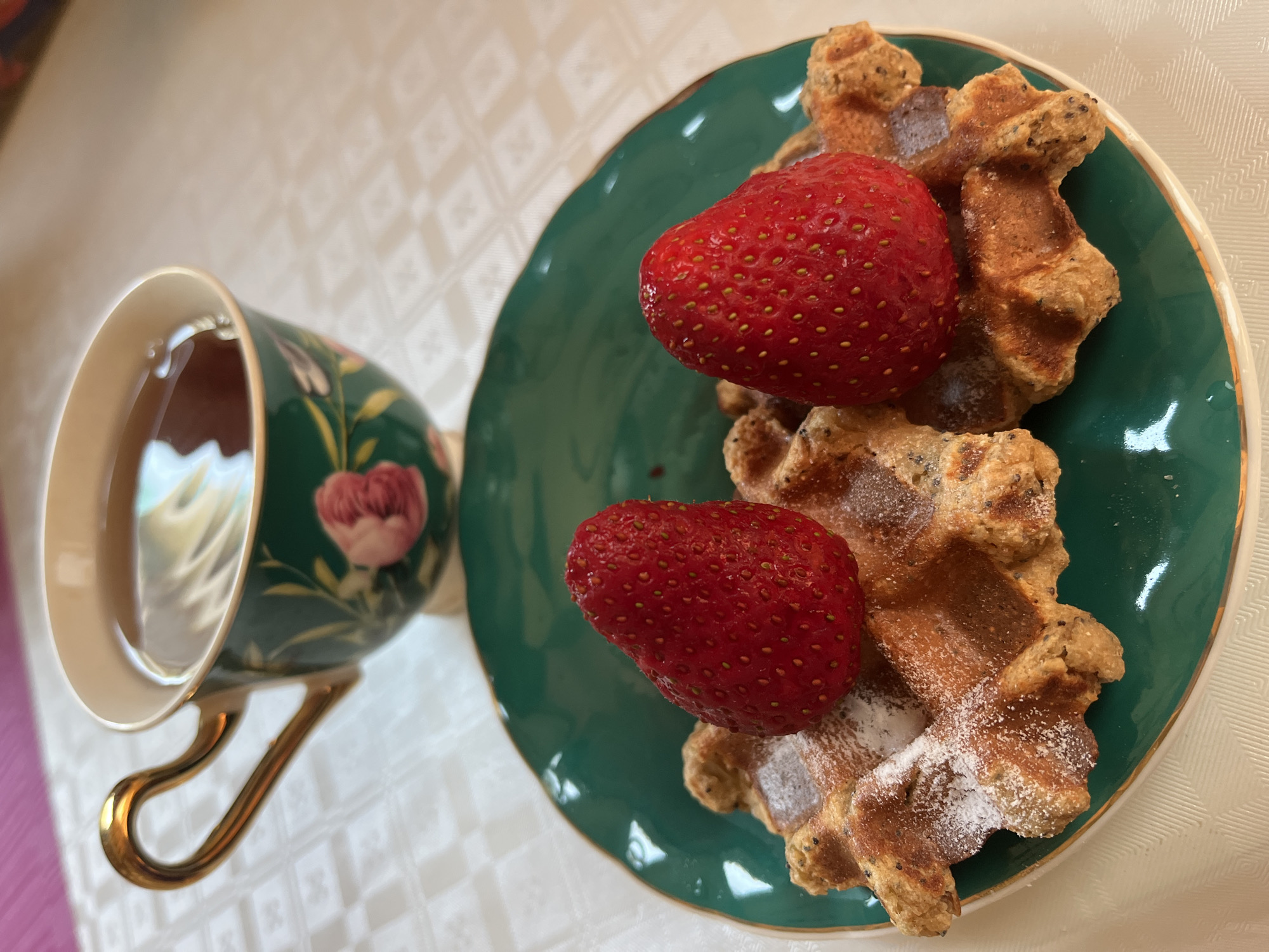 A high-angle, close-up shot depicts a breakfast scene on a white tablecloth, likely indoors. A teacup, patterned with flowers and filled with tea, stands to the left of a round, green plate. The plate holds two waffles, dusted with powdered sugar, and topped with two strawberries. The location, Alanya, Turkey, is not discernible from the visual information.