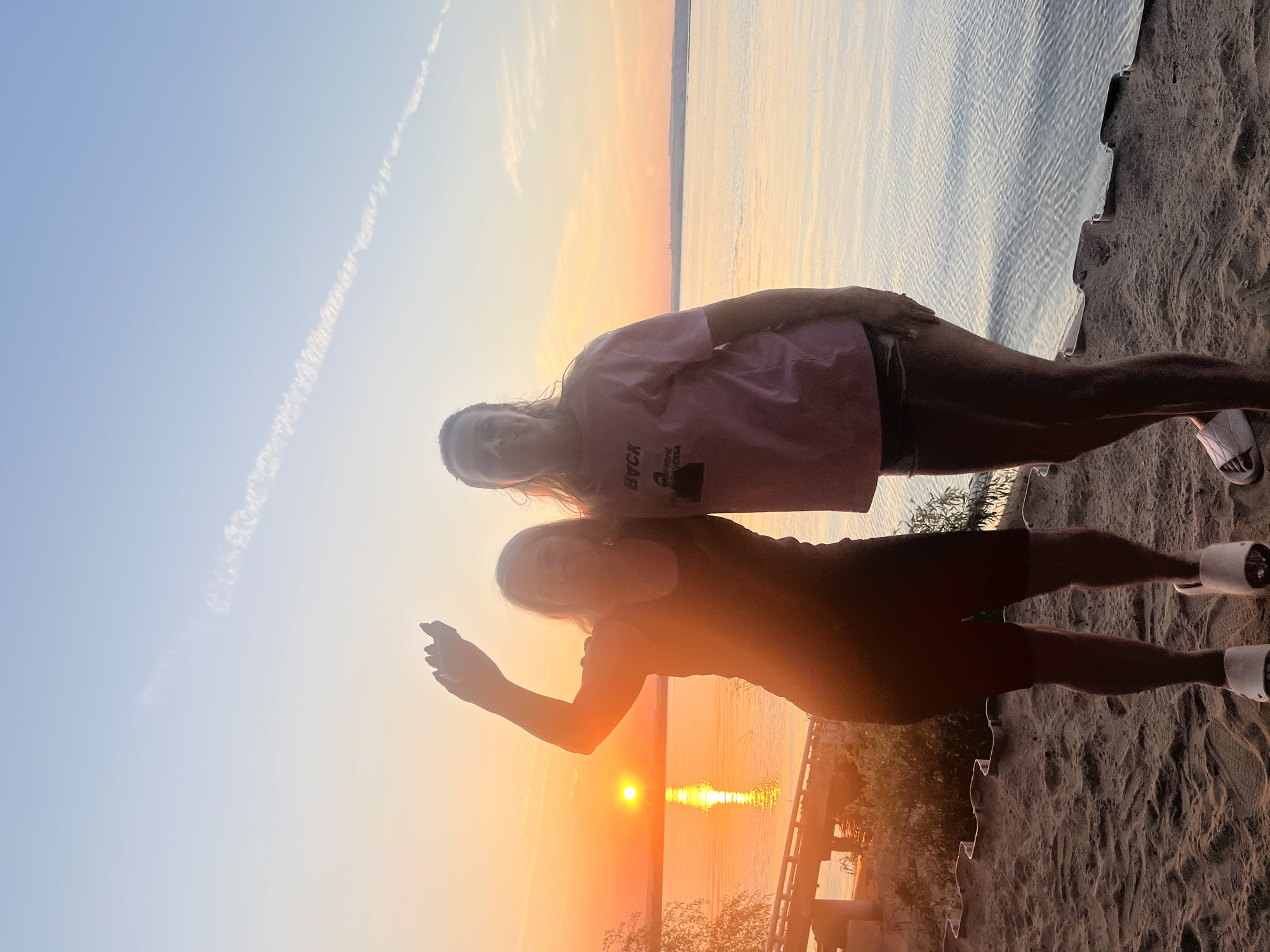 The image captures two women standing on a sandy embankment overlooking a body of water at sunset. The sun, a bright orange orb, is low on the horizon, casting a warm glow across the scene. The sky is a gradient of soft blues and oranges, with a thin trail of clouds stretching across it.
The woman on the left is wearing a dark tank top and dark shorts, with white slides on her feet. She is raising her right hand, as if waving or reaching out. The woman on the right is wearing a pink t-shirt with text on it and dark shorts, also with white slides. She is standing with her arms at her sides. They appear to be casually posing for a photograph.
The foreground consists of coarse sand, with a low, decorative border made of metal or plastic. Behind the women, the water stretches out, reflecting the colors of the sunset. In the distance, the shoreline is visible, with some foliage and a structure that appears to be a bridge or pier on the left side of the image.
The setting suggests a natural, outdoor environment, likely a beach or a riverbank. Given the context of Shumeika, Russia, this could be a waterfront area within or near the city, utilized for recreation during the warmer months. The lighting indicates it is late afternoon or early evening.