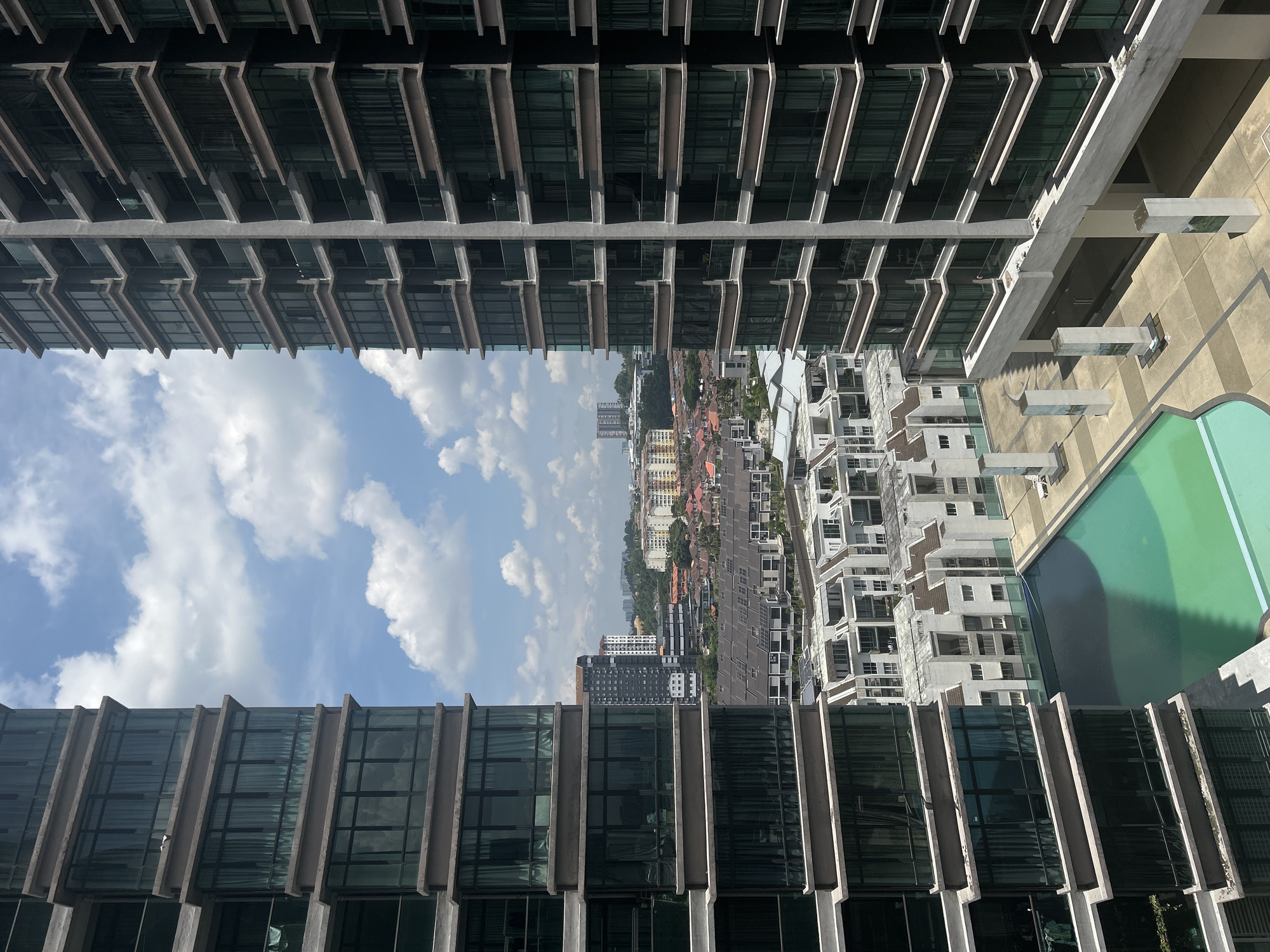 The image shows a view of Kuala Lumpur, Malaysia, seen through a gap between two buildings. The buildings have a modern architectural style with glass windows and concrete supports. A swimming pool is visible on the right side of the image. In the distance, residential buildings and the city skyline are visible under a cloudy sky.