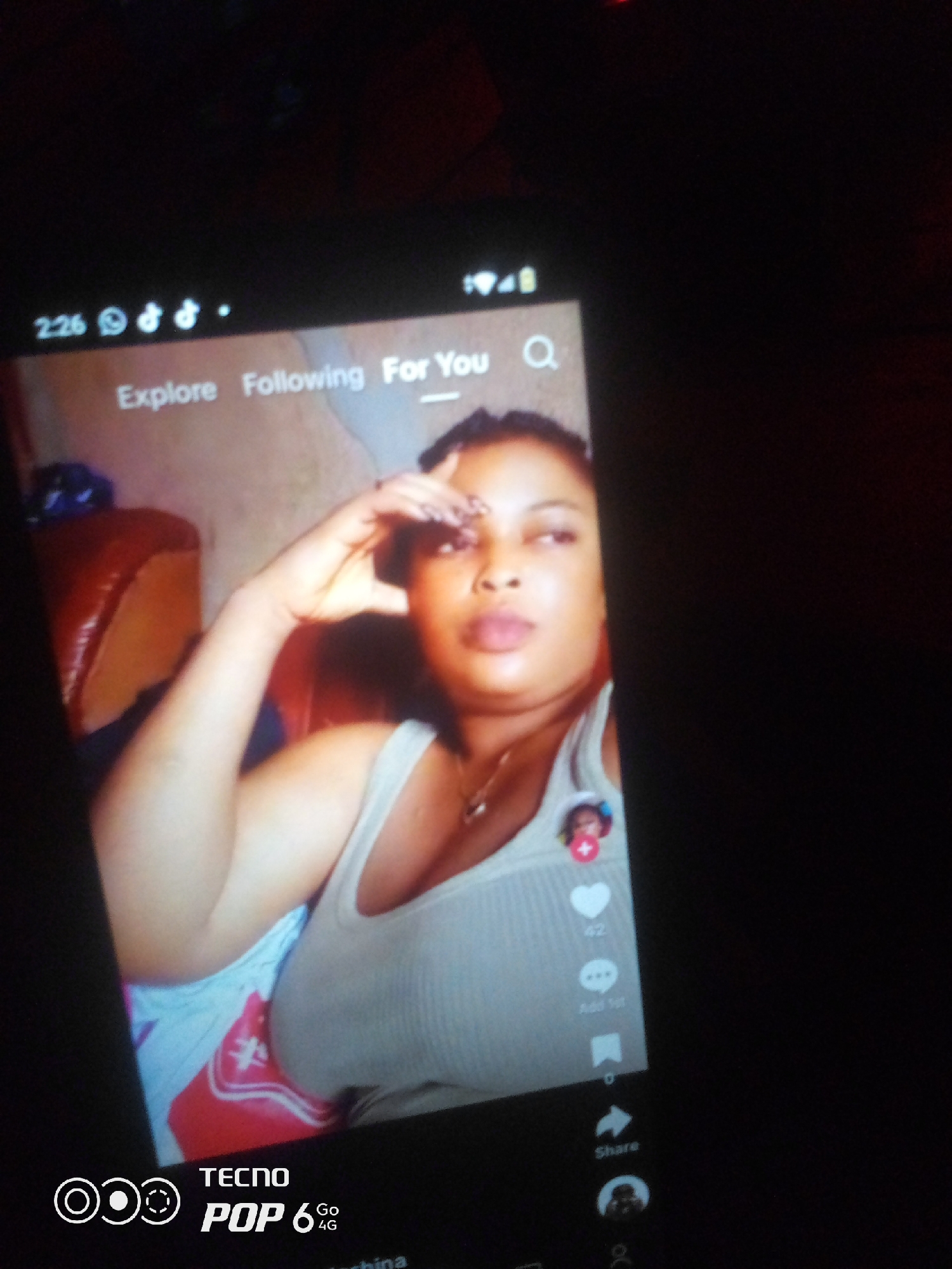 The image shows a close-up of a smartphone screen displaying a social media video. The video features a woman with dark hair and makeup, wearing a light-colored tank top. She is looking towards the viewer and has her hand on her forehead. On the right side of the screen, there are interactive elements like a heart icon with the number "42" next to it, a speech bubble icon, and a share icon. The phone's interface displays "Explore," "Following," and "For You" at the top. The bottom of the screen shows the "TECNO POP 6 Go 4G" branding. The background of the video appears to be an indoor setting with a brown leather chair visible on the left. The overall lighting suggests a dimly lit environment. This image does not provide any information about the location in the city of Howayi, Nigeria.