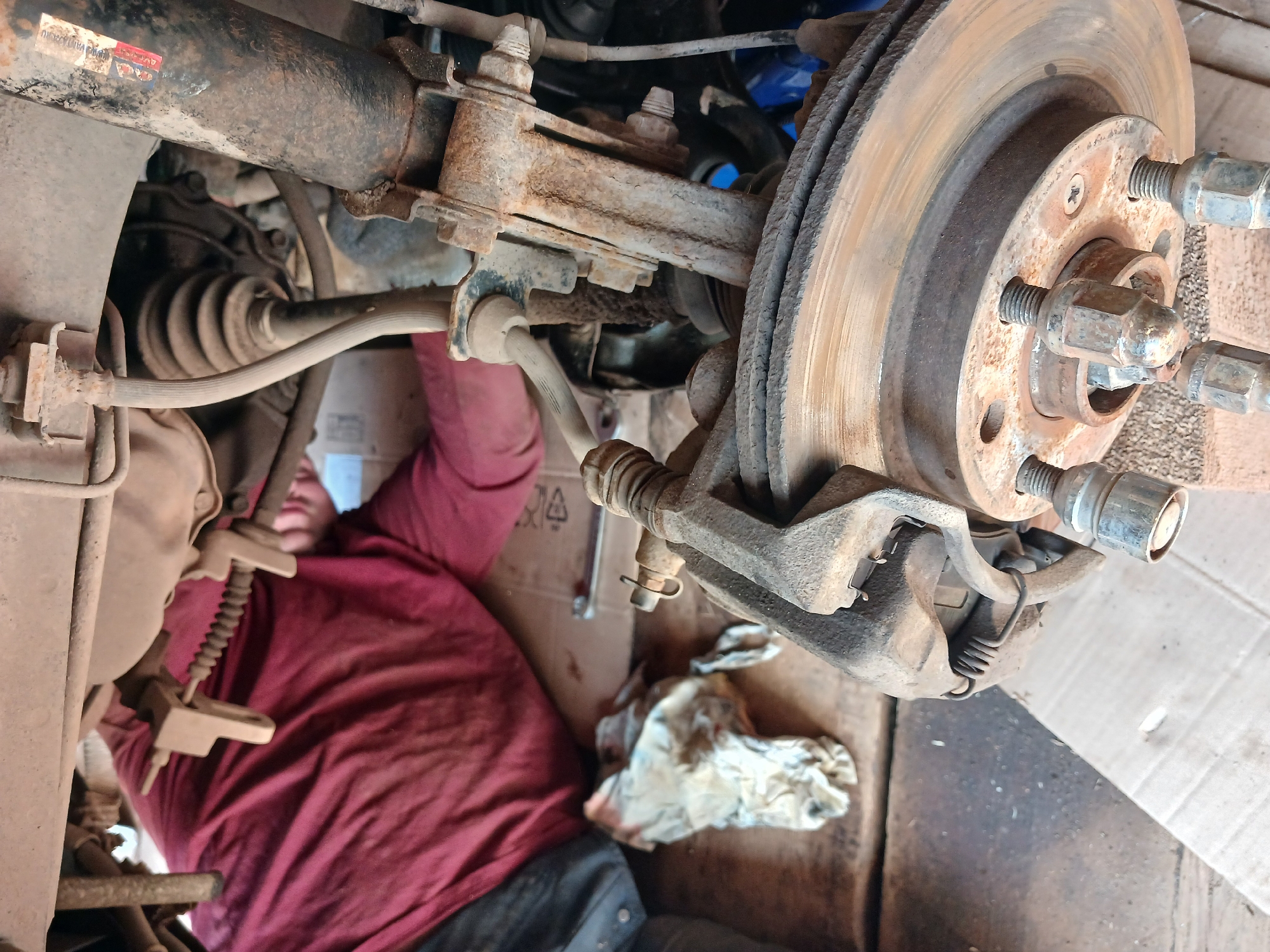 A person is lying on their back, working on a vehicle. The vehicle's wheel has been removed, exposing the brake rotor, caliper, and suspension components. The brake rotor is rusty, and the surrounding parts are heavily weathered and show signs of wear. The person is wearing a maroon shirt and dark pants. A dirty rag and tools are near the person. The scene is cluttered and appears to be a makeshift repair area, with cardboard on the floor. The location is Noviy, Russia.