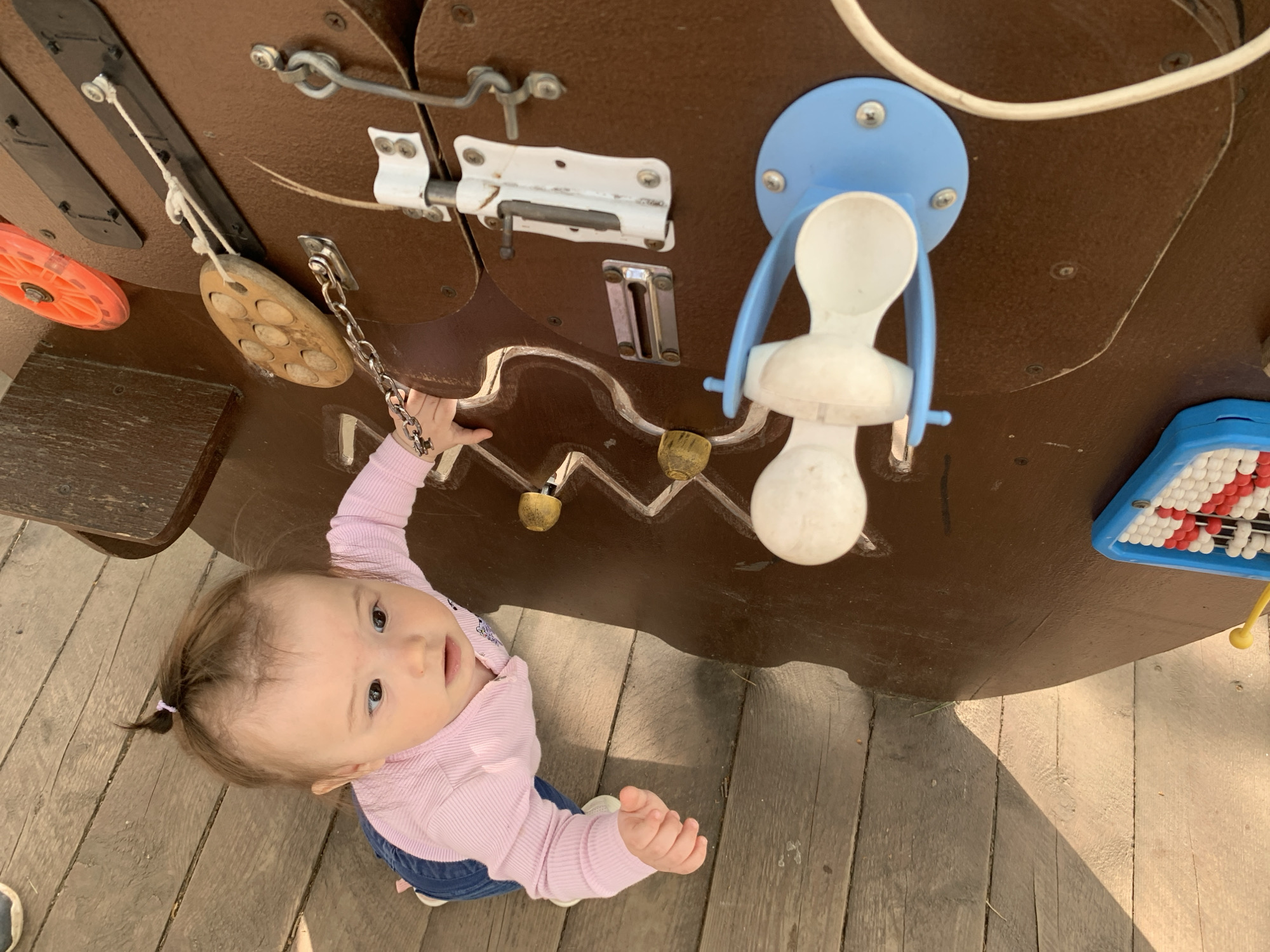 A baby girl, likely between 9-15 months old, is reaching up to interact with a wooden activity board mounted on a wooden structure. The board features various mechanical elements including latches, wheels, knobs and a chain. The baby is wearing a pink long-sleeved shirt and jeans. The wooden structure appears to be part of a playground or outdoor play area. The scene is located outdoors in Chaikovsky, Russia.