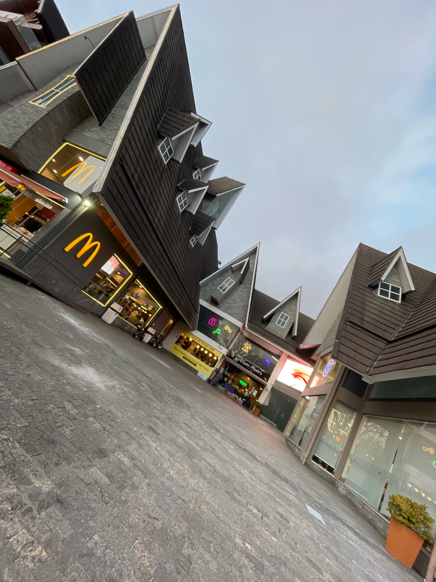 The image captures a street-level view of a commercial area in Gramado, Brazil, characterized by buildings with unique architectural styles, featuring steep, dark brown roofs reminiscent of European chalets. The buildings are adorned with prominent signage, including a large, illuminated McDonald's logo on the left. Storefronts with large glass windows line the street, with some showing interior lights and displays. The pavement is made of cobblestones. The sky is overcast and appears to be late afternoon or early evening, indicated by the artificial lighting illuminating the storefronts. There are a few people visible, but their interactions are not discernible. The overall scene suggests a tourist-oriented commercial district, possibly with themed architecture to attract visitors.