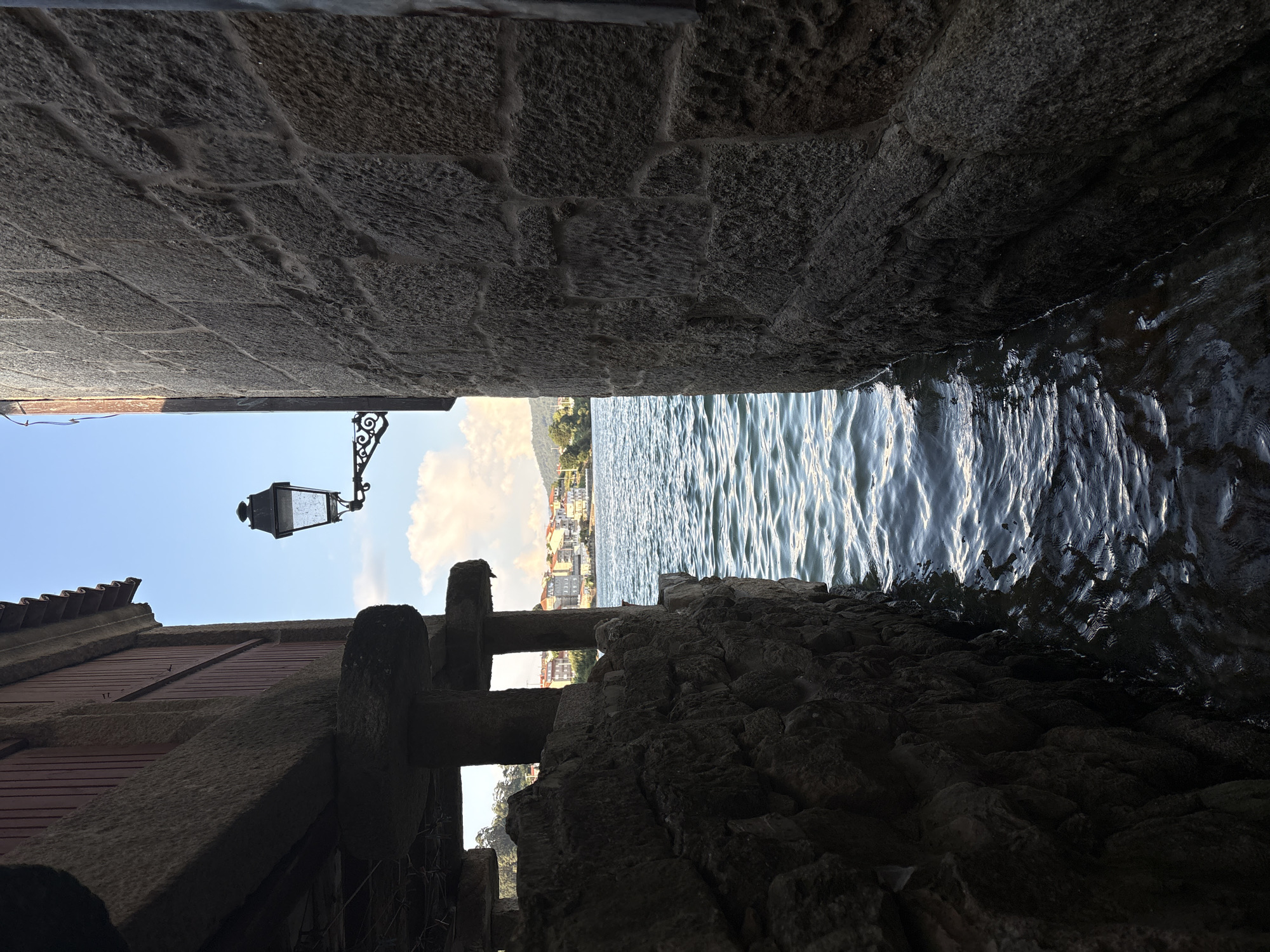 The image shows a narrow waterway between two stone buildings, with the sea visible in the background. On the left, a building with wooden siding and a red roof is supported by stone pillars. A lamppost with an ornate bracket is mounted on the wall of the building on the right, which is constructed of rough-hewn stone blocks. The water is choppy and reflects the sunlight. In the distance, across the water, is a town with buildings and a green hillside. The scene is located in the town of Combarro, Spain, a coastal village known for its traditional architecture and seafood restaurants. The time of day appears to be late afternoon or early evening, as indicated by the warm sunlight and the shadows.